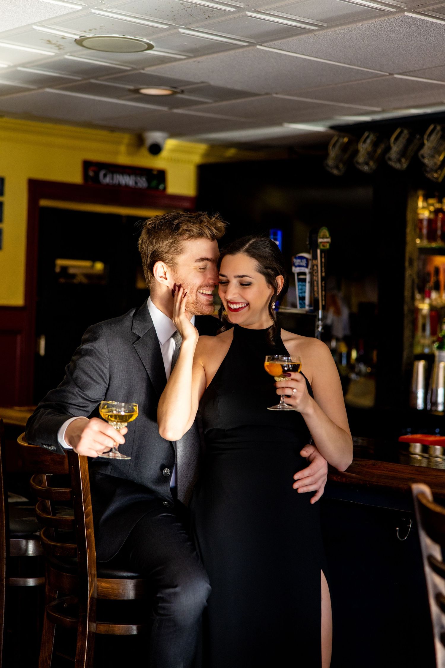 Elegantly dressed couple sharing drinks and laughter at a bar counter.