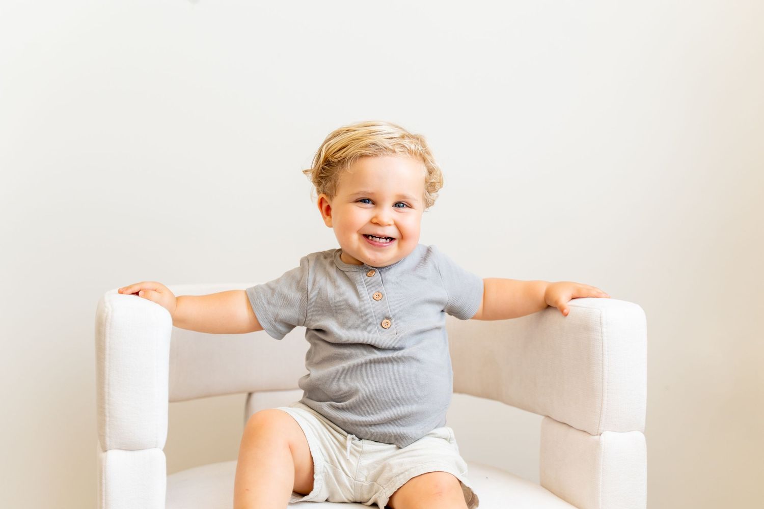 Happy toddler sitting on white chair against neutral background.