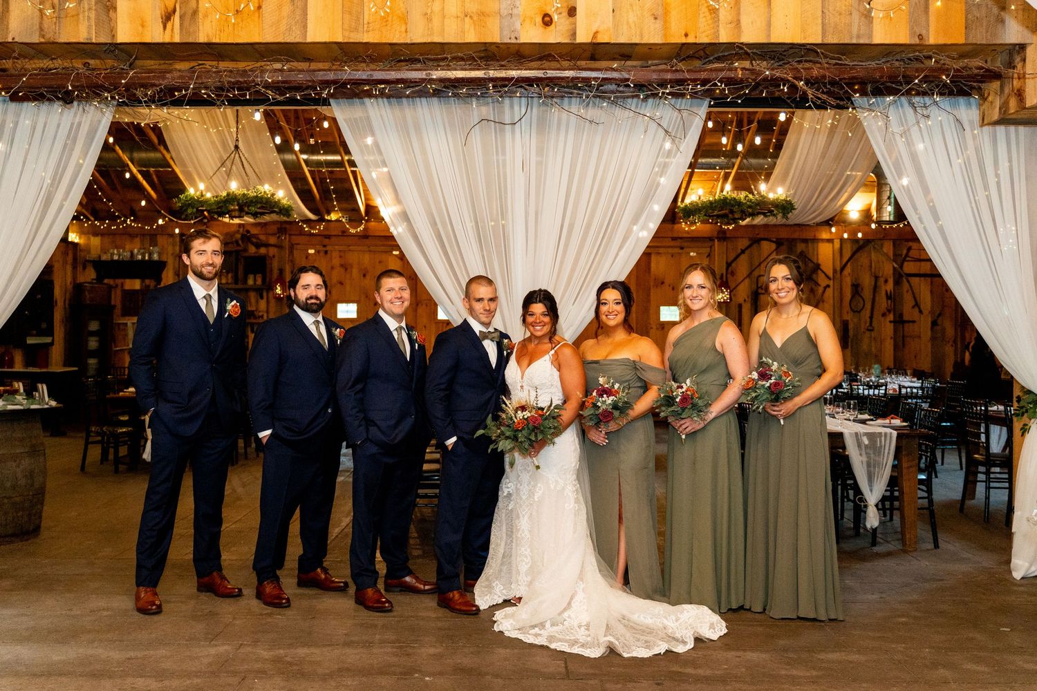 Wedding party lined up in rustic barn venue with white draping and wooden beams.