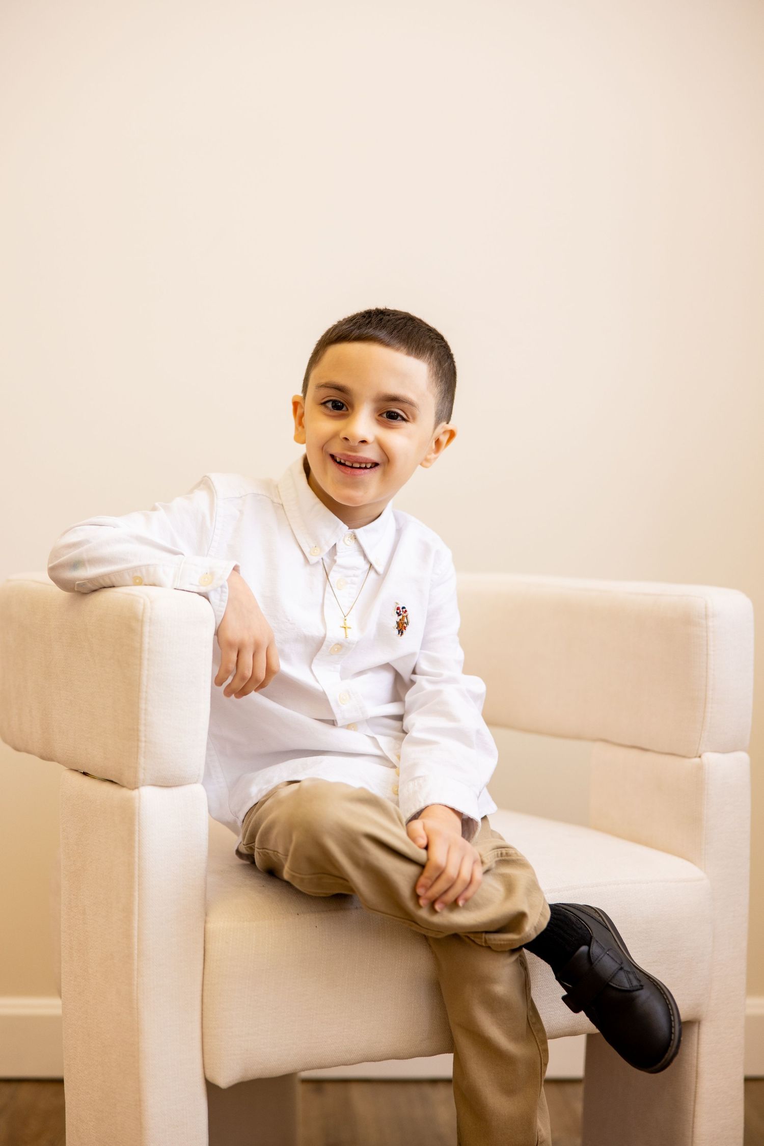 A formal portrait of a young person in white dress shirt and khakis sitting on beige chair.