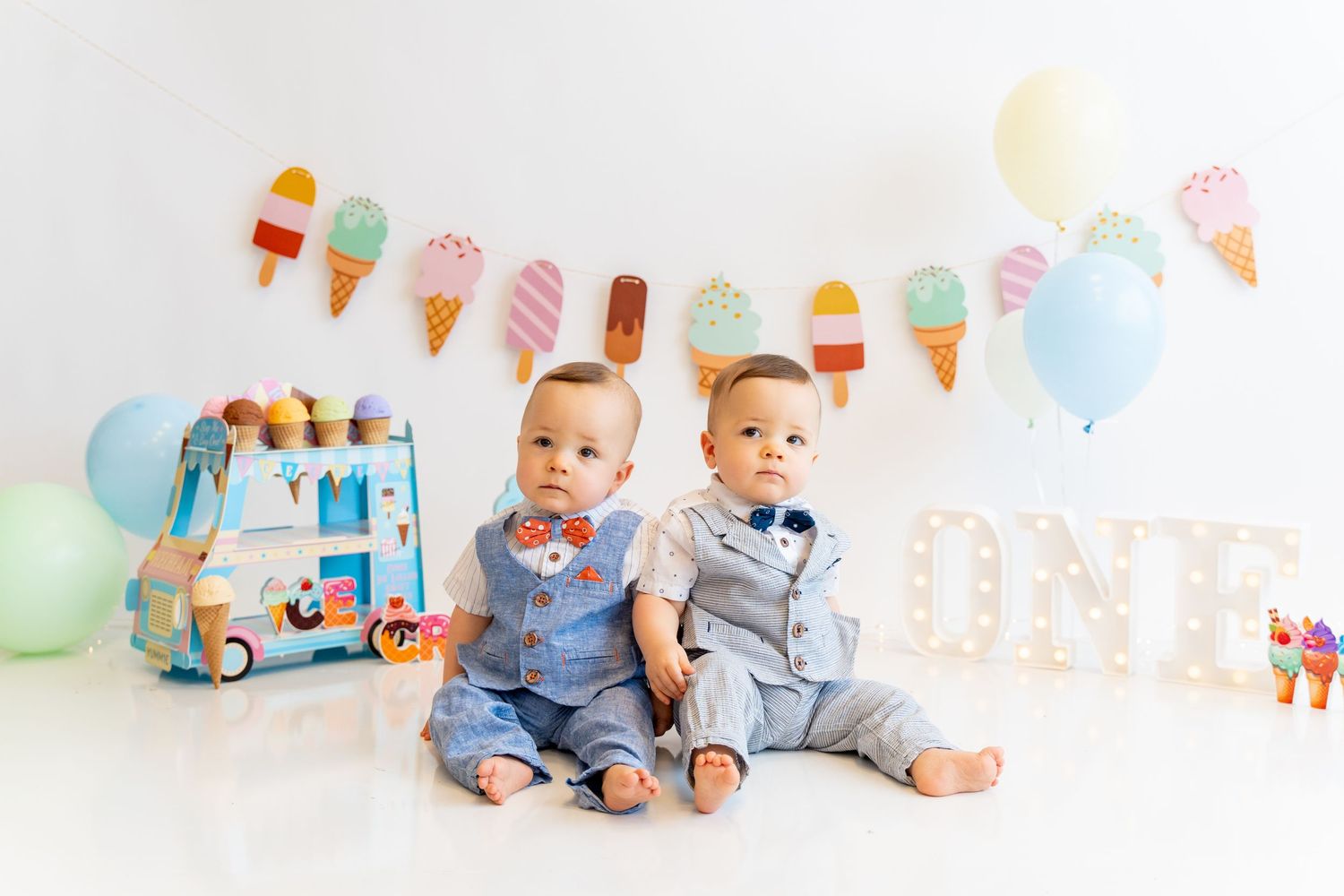 Two babies in coordinating blue outfits sitting against ice cream themed party backdrop.