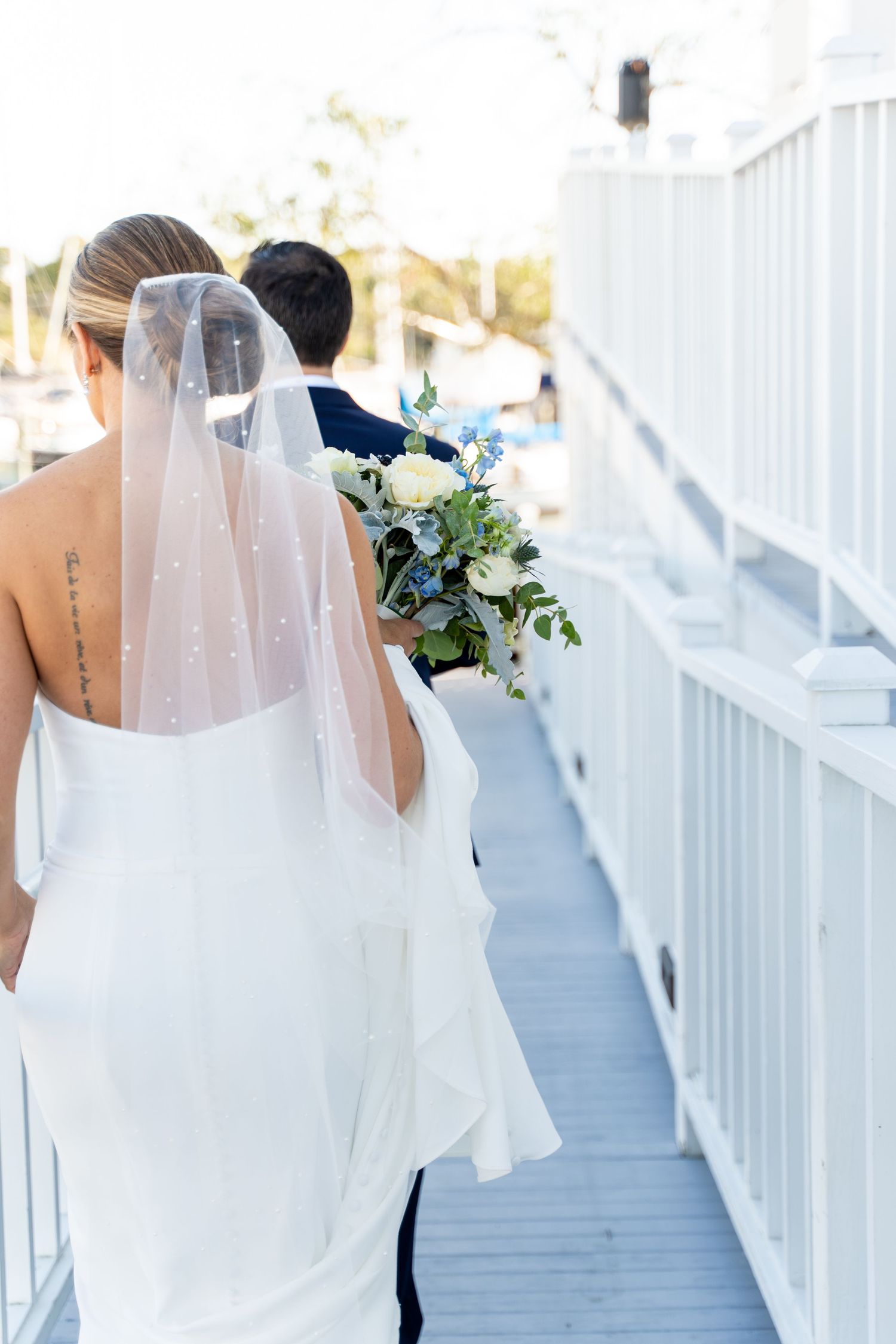 Bride in white dress with flowing veil walking along waterfront boardwalk.