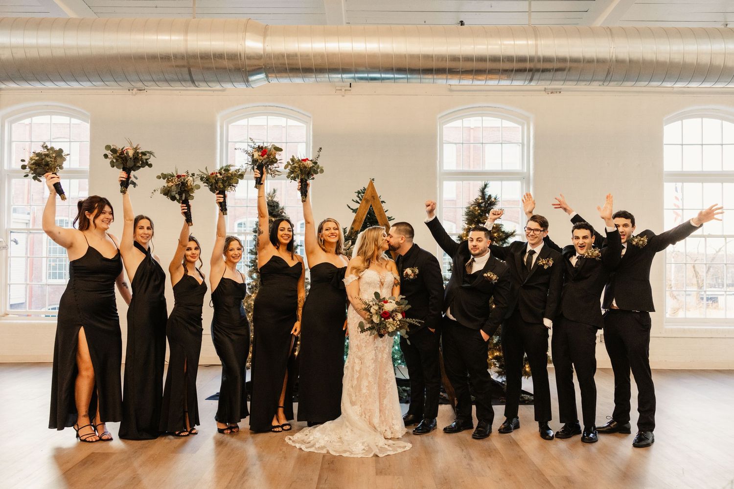 Joyful wedding party celebrates with raised bouquets in bright industrial venue.