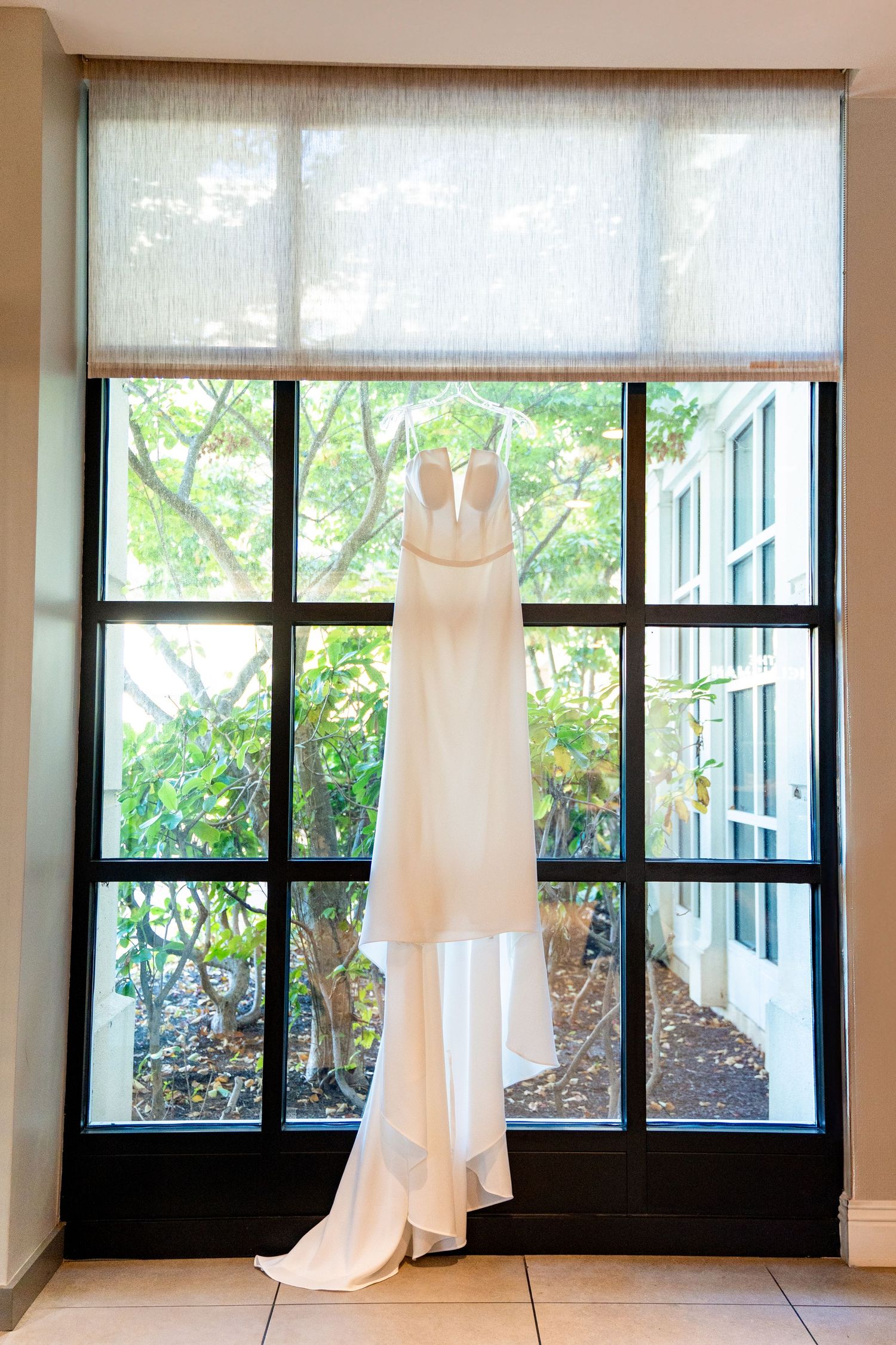Elegant white wedding dress hanging in front of a large window with garden view.