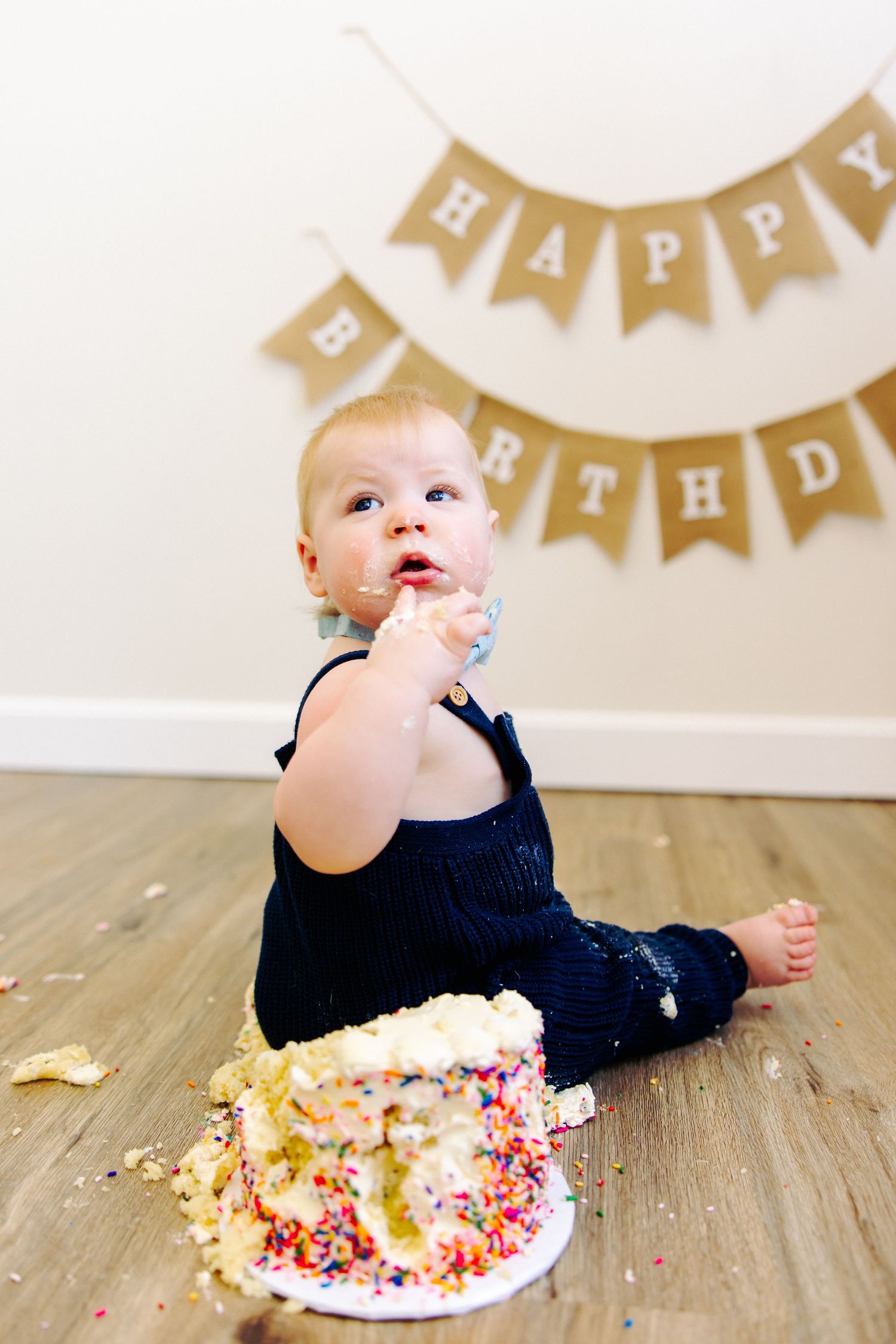 Baby in navy outfit tastes frosting while sitting next to smashed birthday cake.