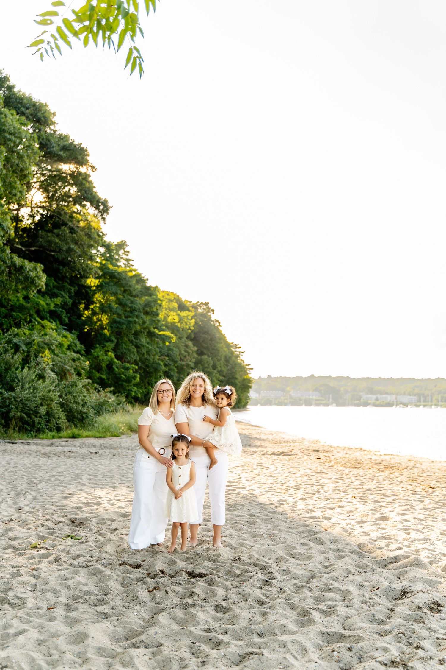 Family group in white outfits poses on sandy beach with forest backdrop at sunset.