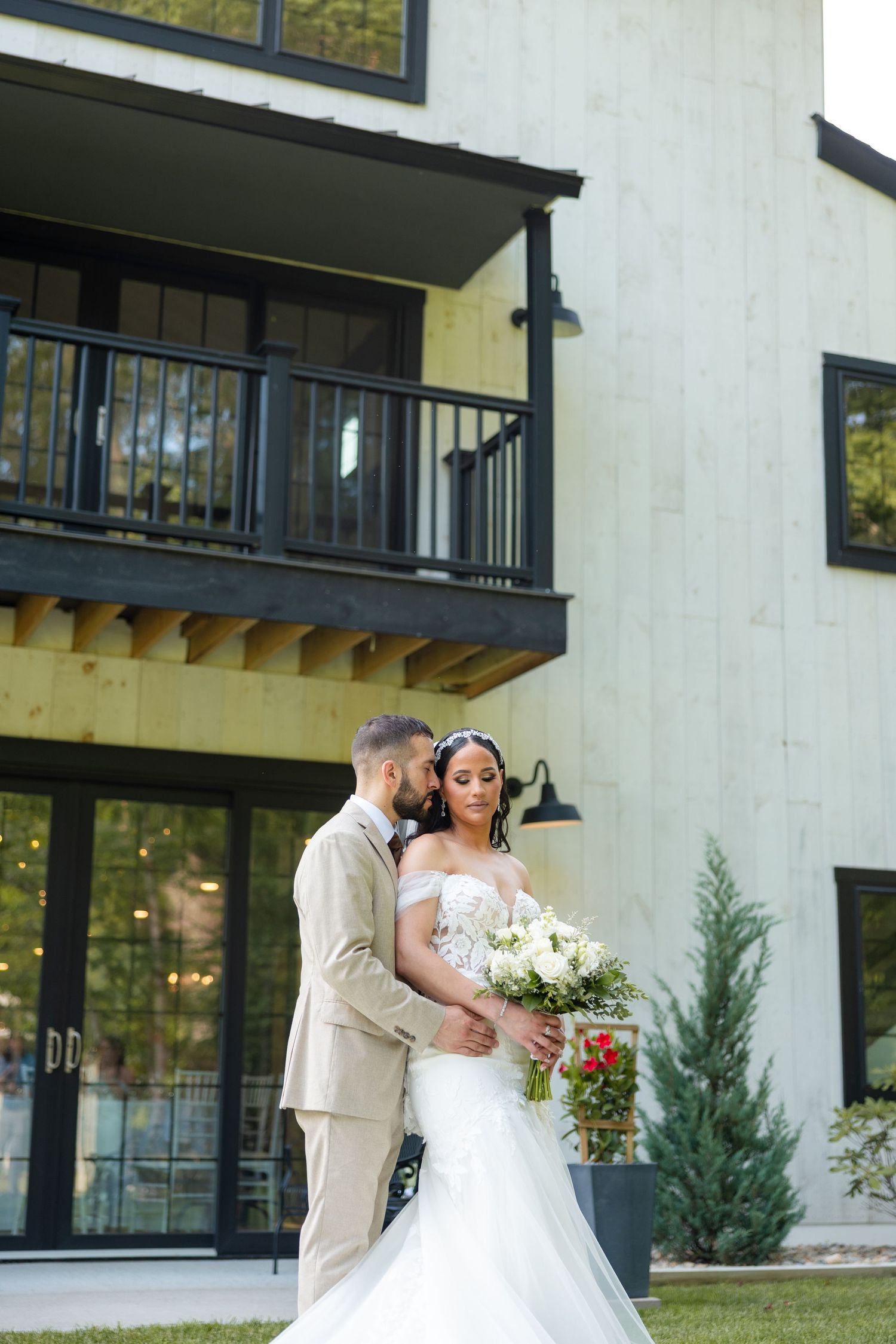 A bride in a white mermaid dress holds a bouquet while standing with her partner in a tan suit outside a modern building.
