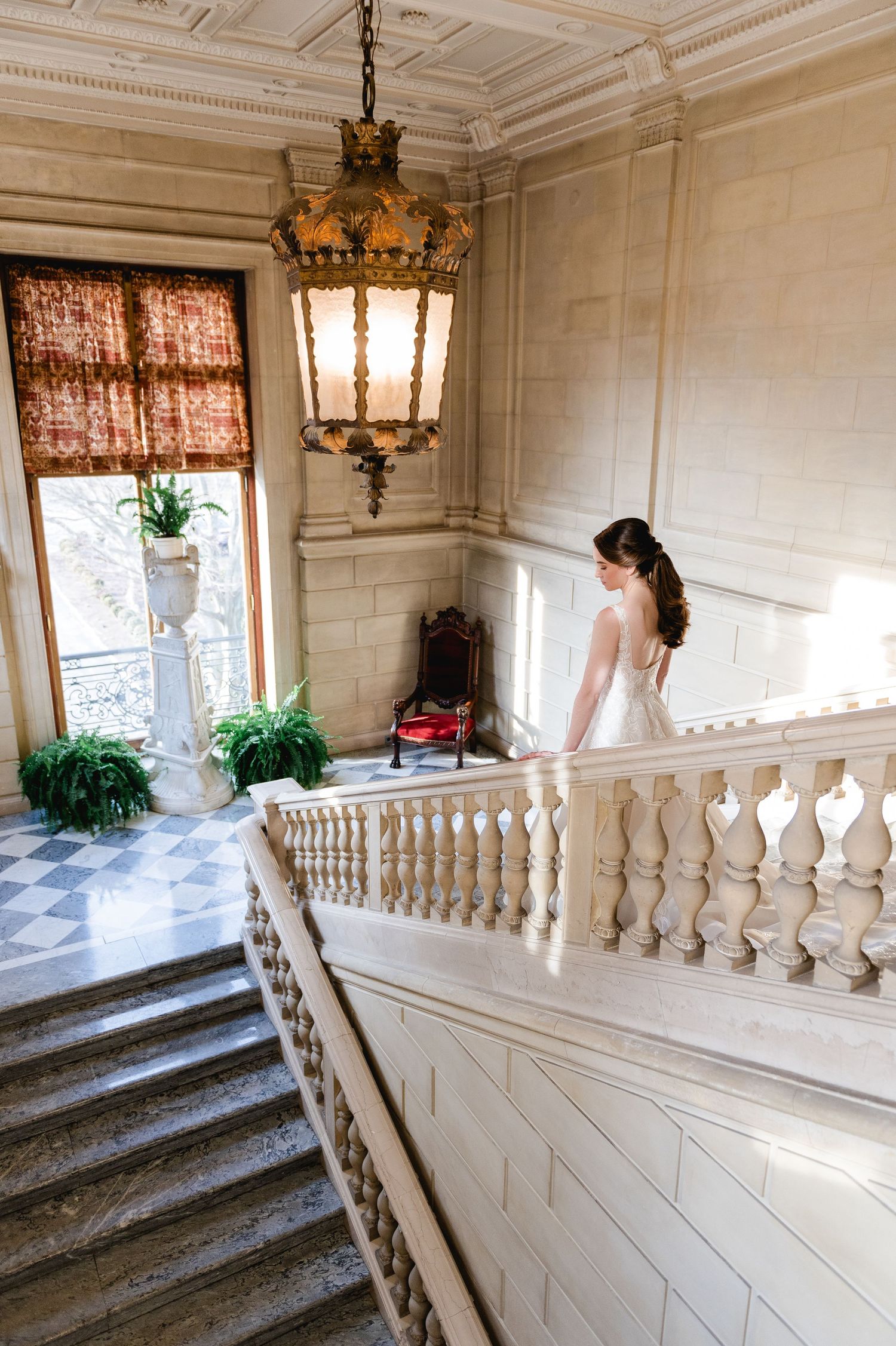 A bride in a white gown stands on an elegant marble staircase with ornate balustrade and vintage lighting.