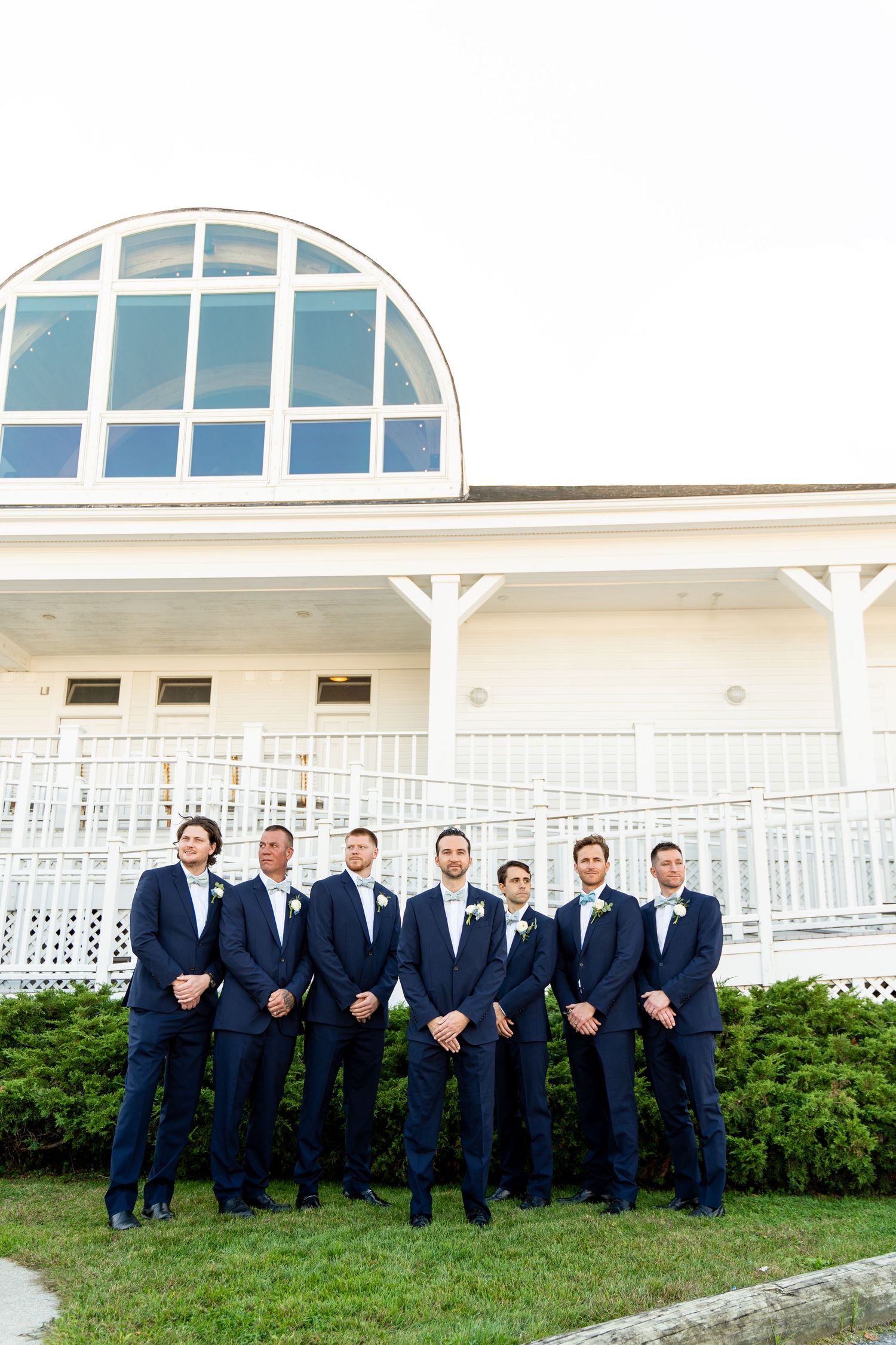Groomsmen in navy suits stand in formation against white building with distinctive arched window.