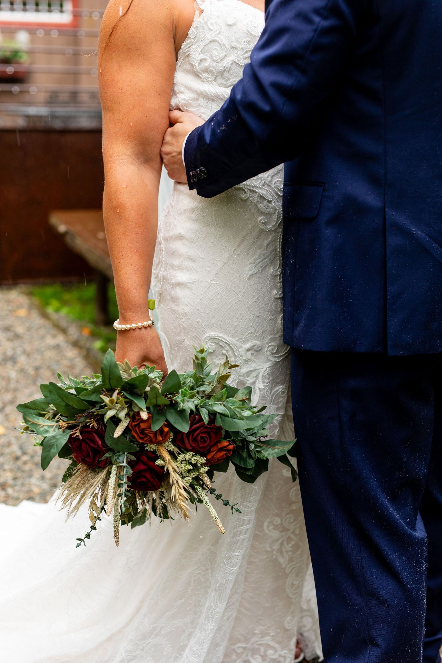 Detail shot of bride holding burgundy and greenery bouquet next to groom in navy suit.
