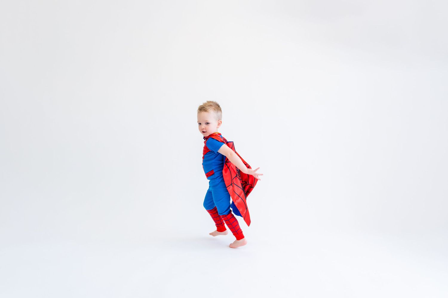 Child in superhero costume with red cape jumping against white background.