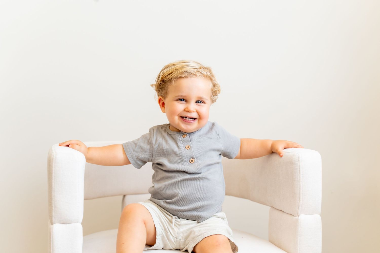 Happy toddler in gray outfit sitting on white chair with arms spread.