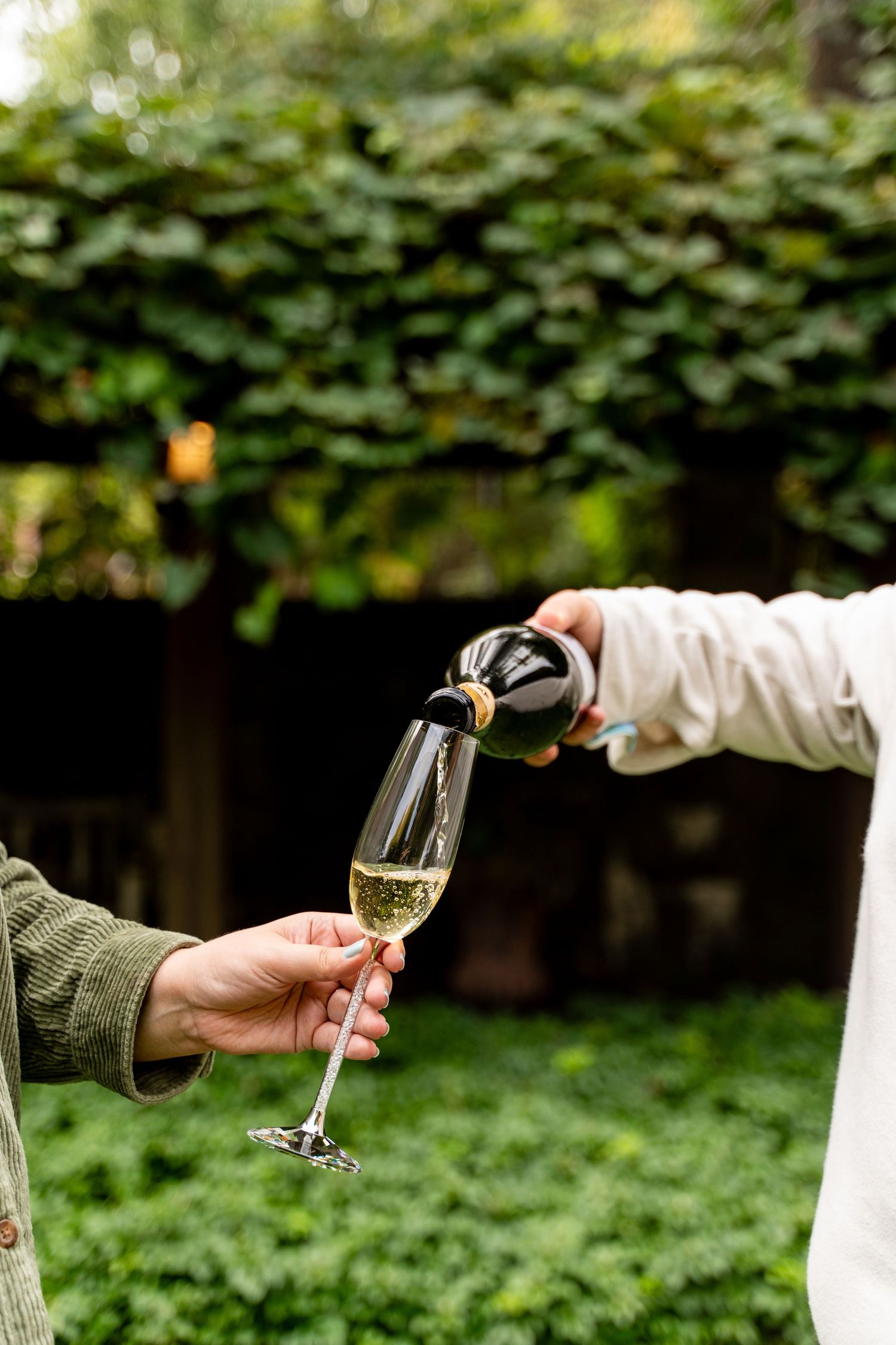 Hands pouring champagne into glass in garden setting.