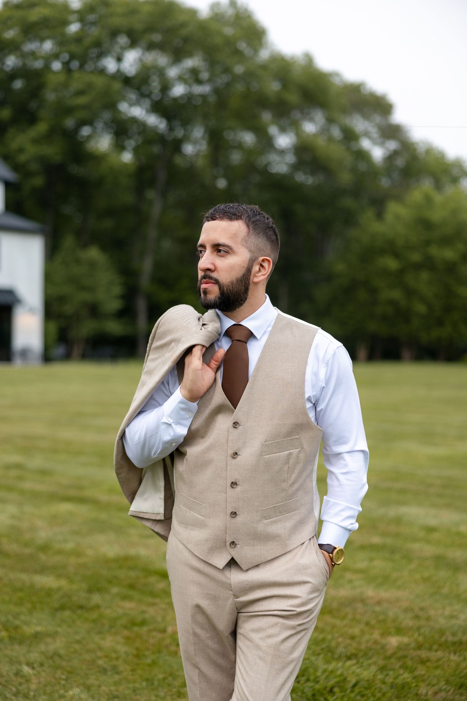 A person wearing a beige vest and white dress shirt poses thoughtfully in a grassy outdoor setting.