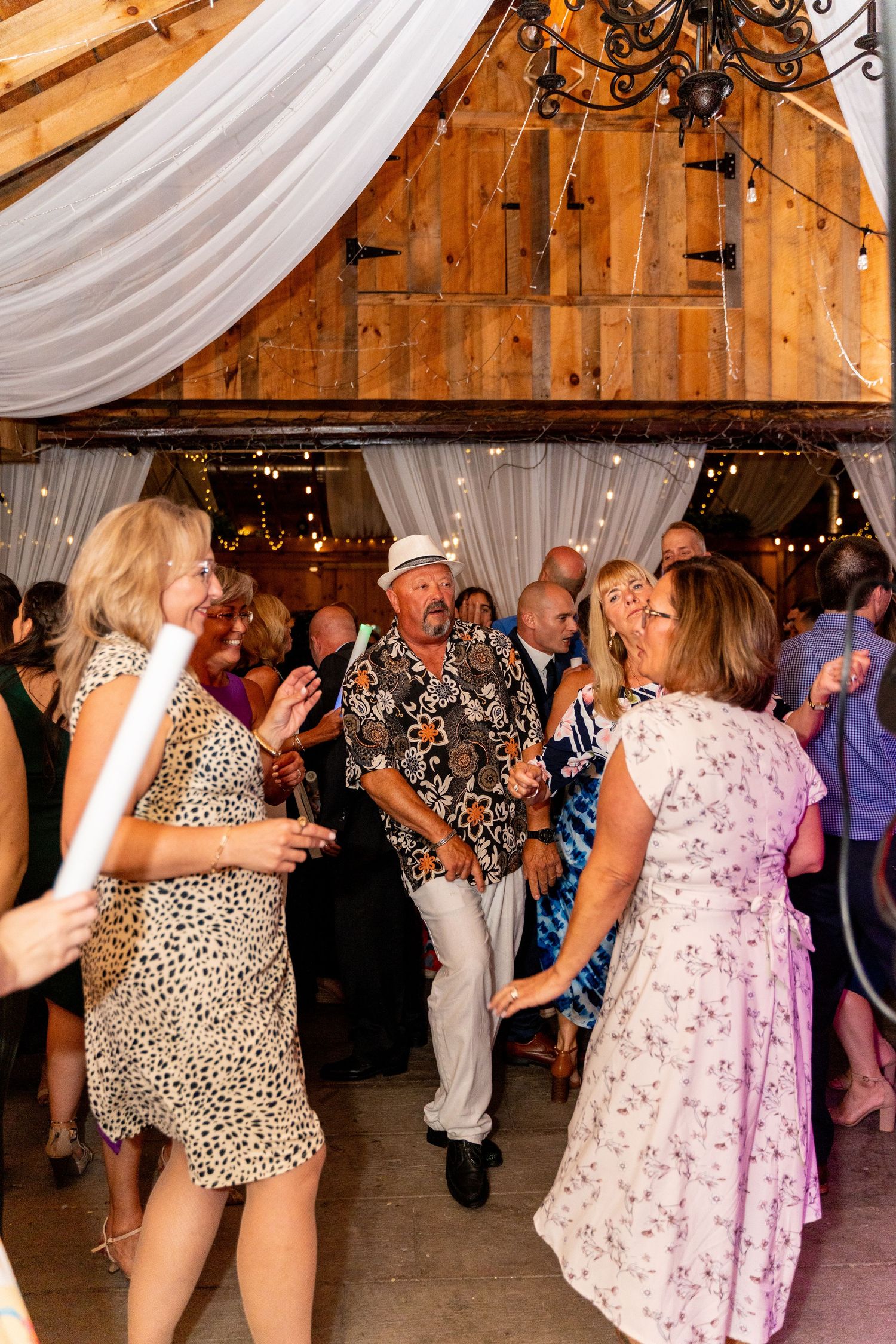 Wedding guests dance and celebrate in a rustic barn decorated with white drapes and string lights.