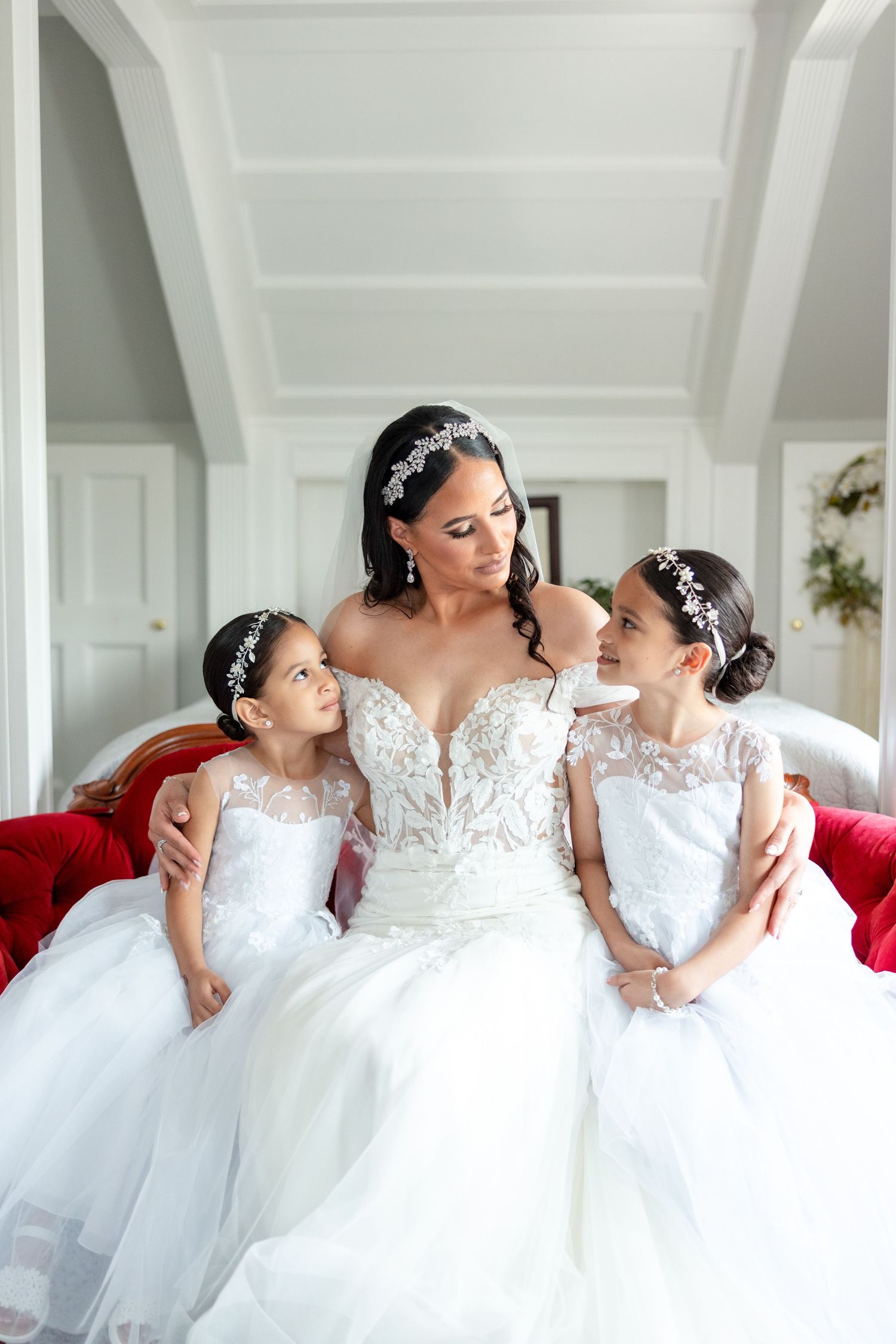 Five bridesmaids wearing white lace wedding gowns pose together on red furniture in an elegant white room.