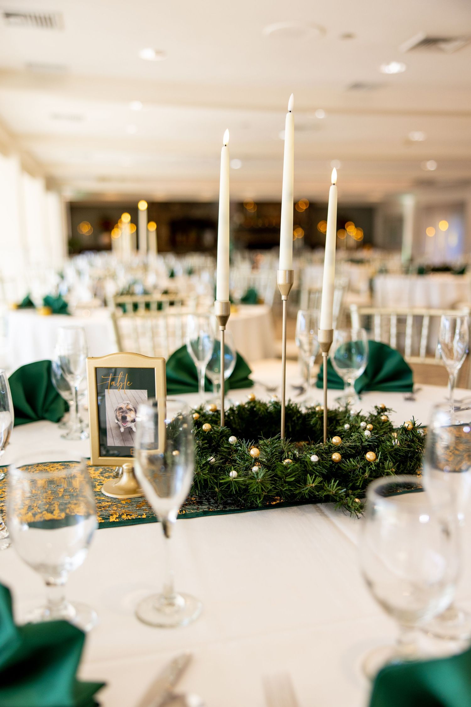 Elegant wedding table setting with white candles, green napkins and evergreen centerpiece with gold accents.