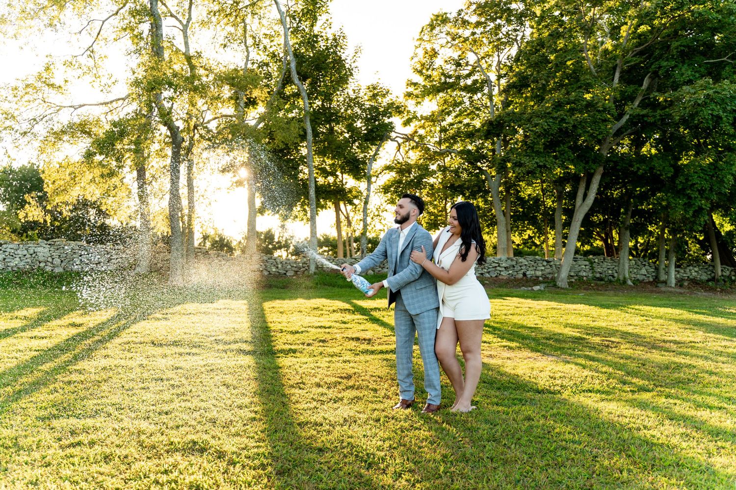 A couple in light-colored formal wear celebrating in golden sunlight on a grassy lawn.