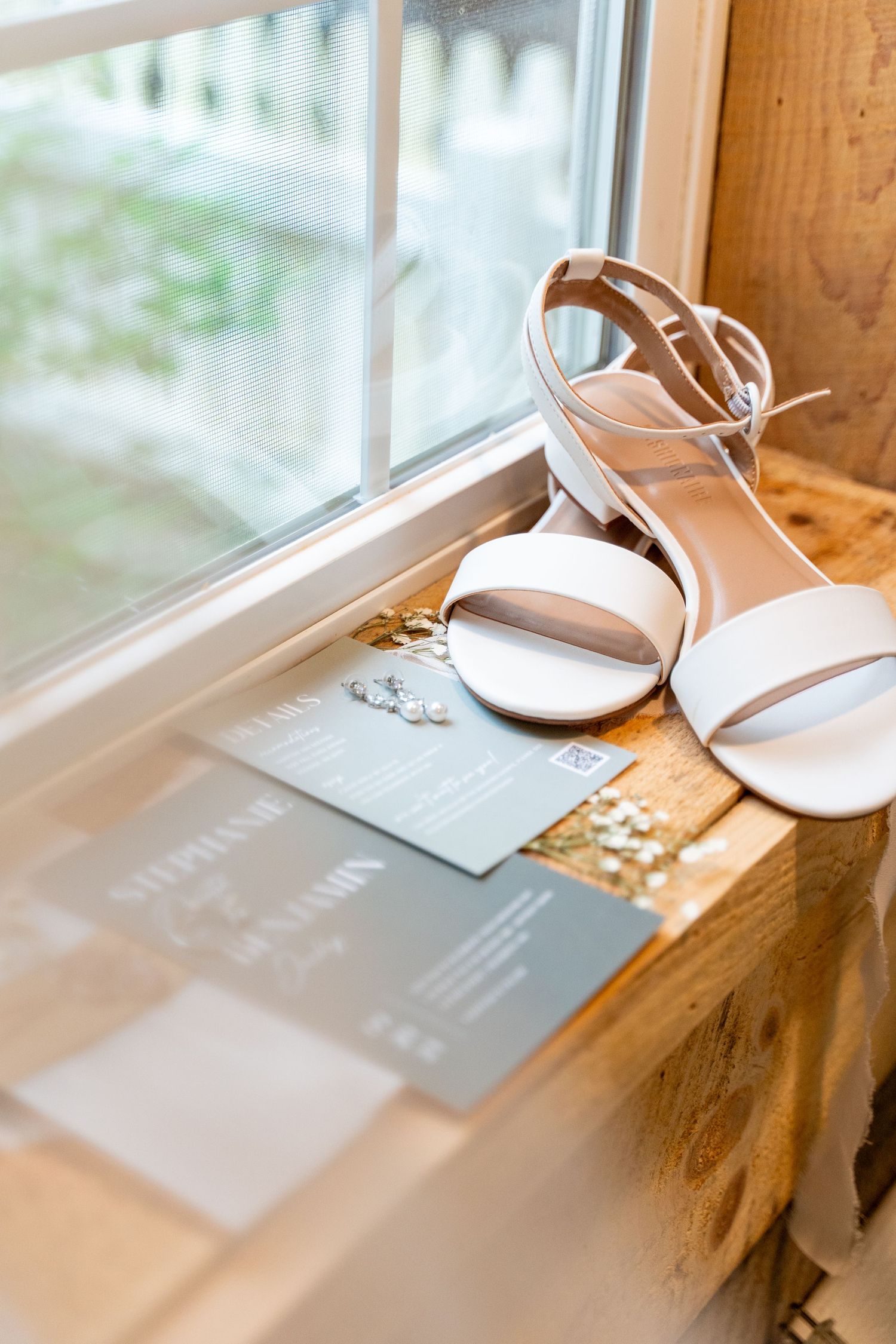 White bridal sandals and wedding invitation displayed on wooden windowsill.