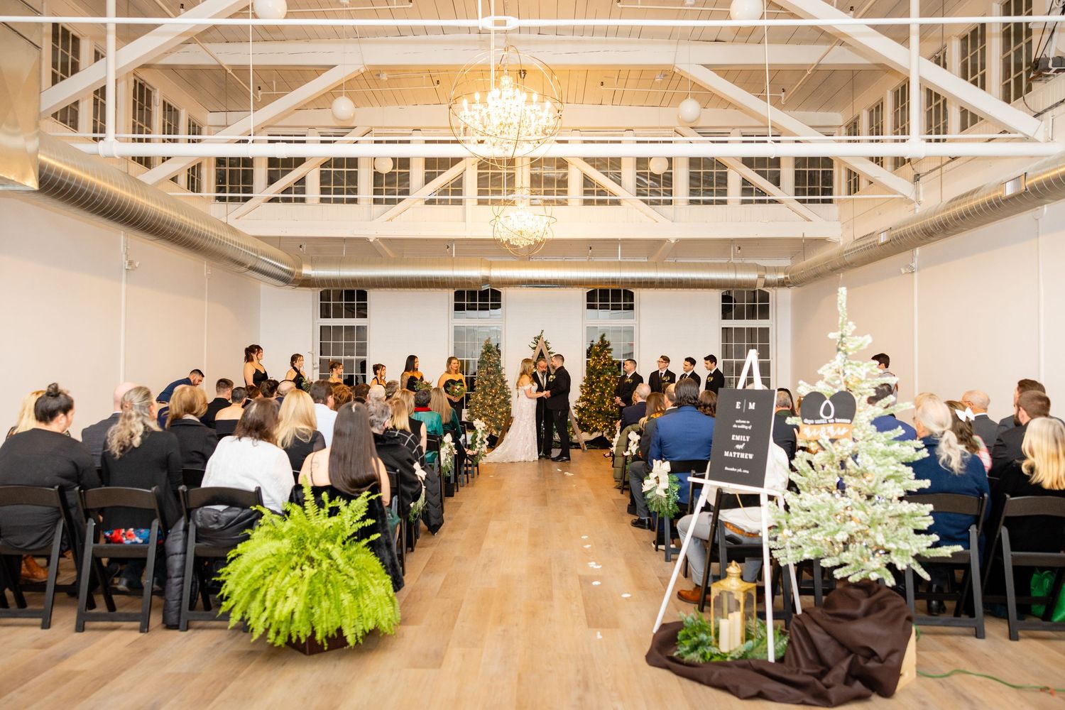 A wedding ceremony setup with black chairs and Christmas trees in an elegant white room.