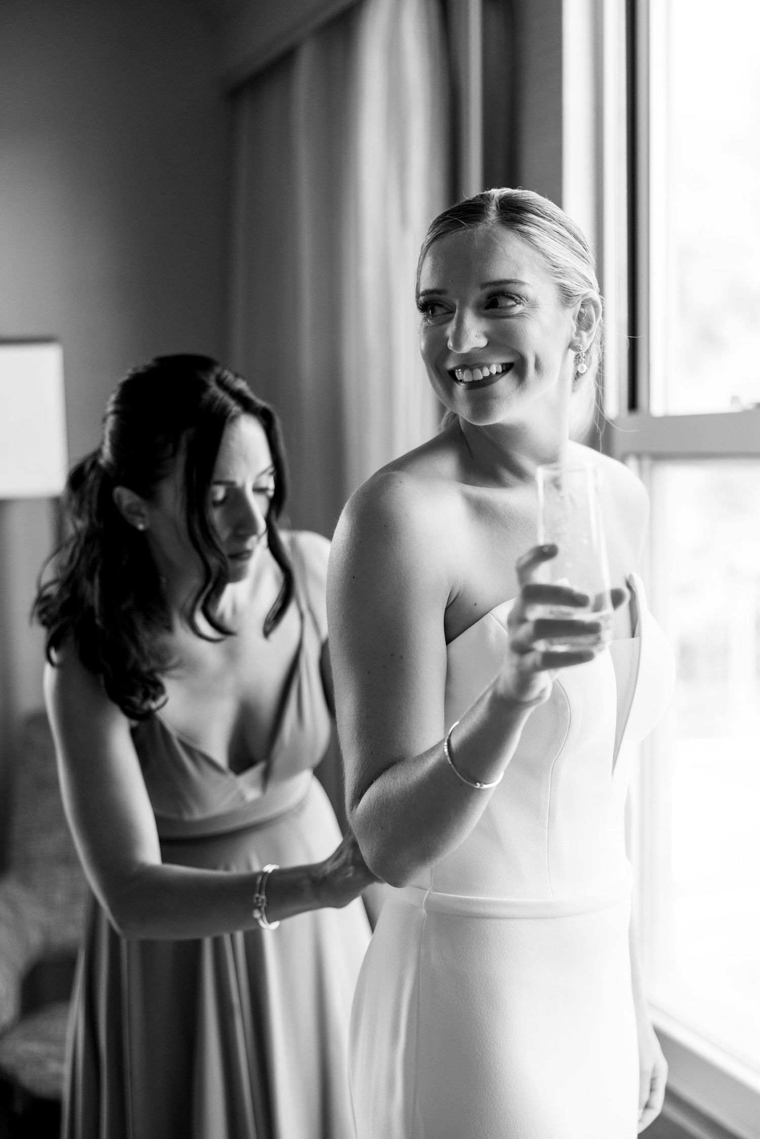 Black and white candid moment of bride and bridesmaid sharing laughter by window.