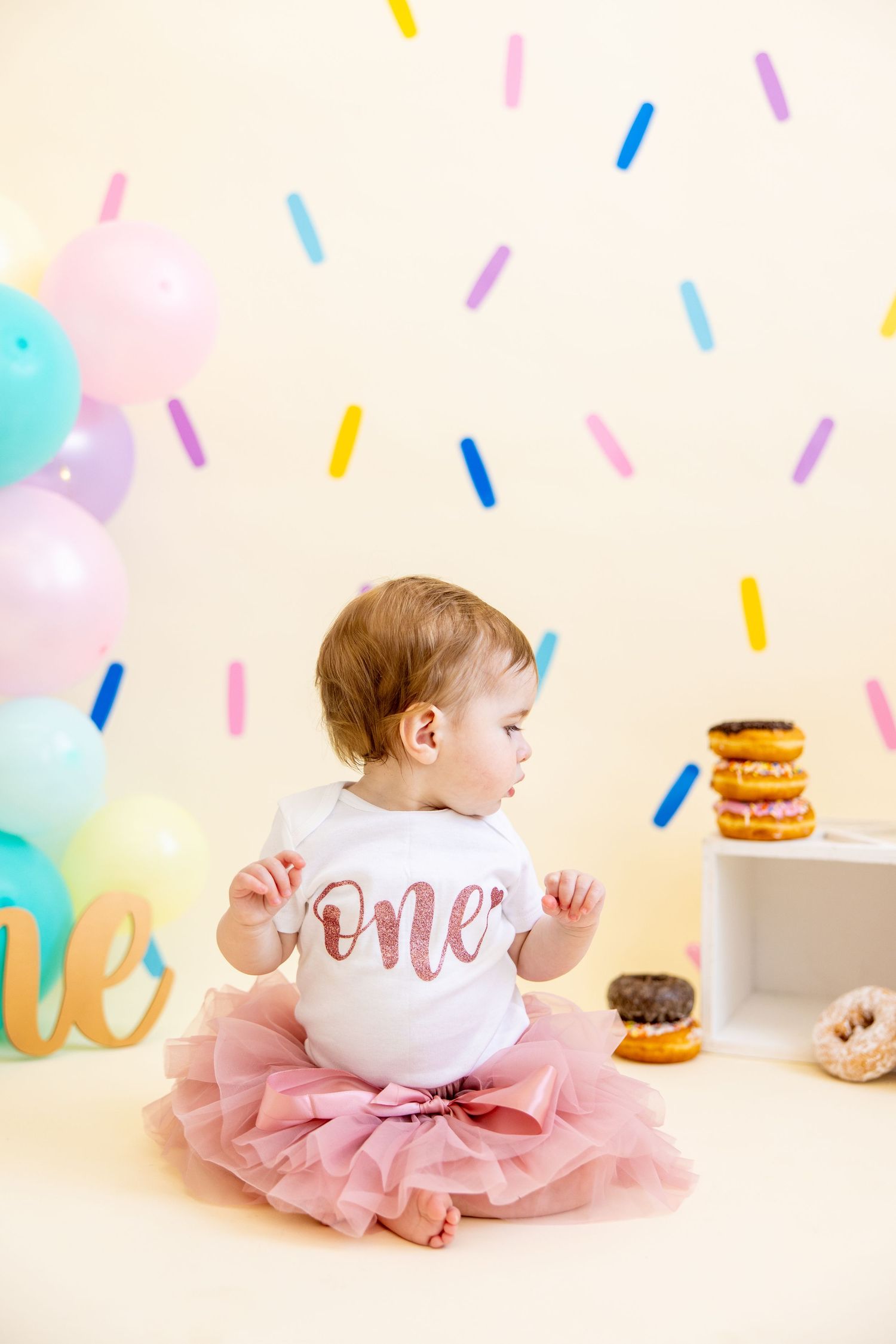Baby in pink tutu looks at donuts while celebrating first birthday with colorful decorations.