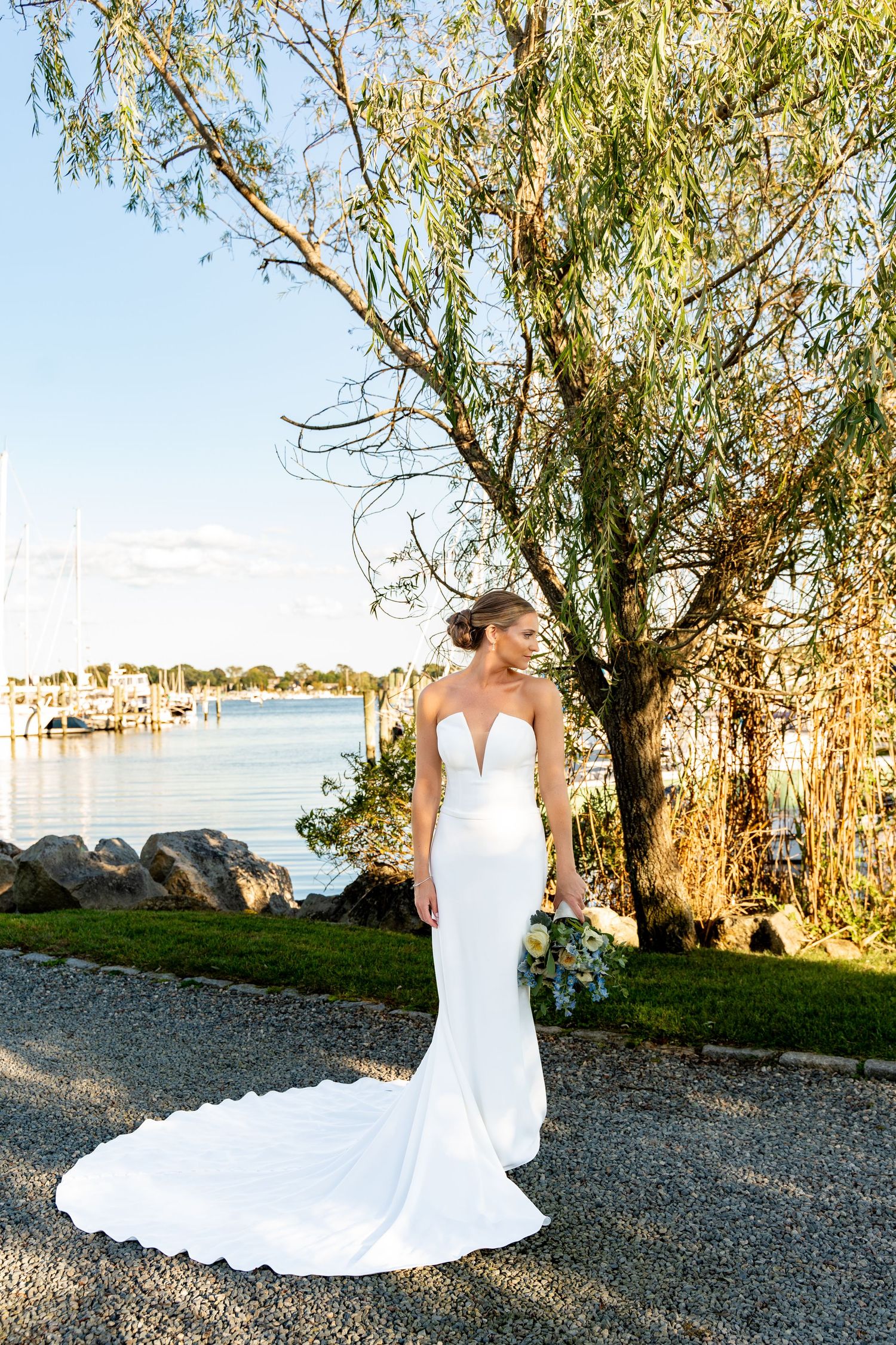 A bride in a fitted white dress with long train stands near a waterfront marina at sunset.