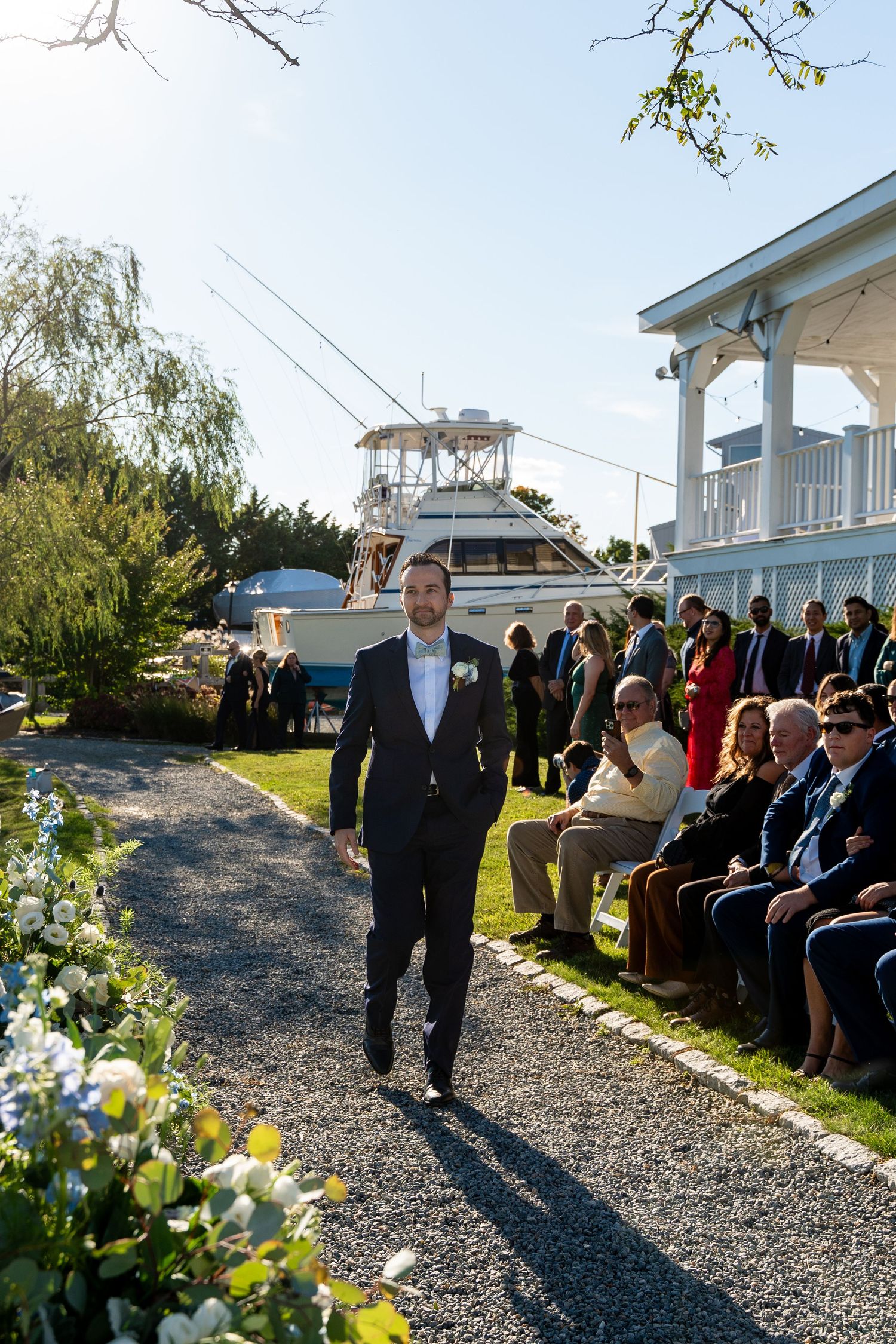 Groom walking down outdoor gravel path with boat and guests at waterfront wedding ceremony.
