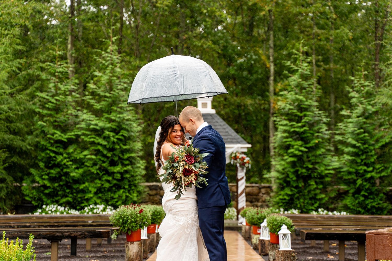 Couple shares romantic moment under clear umbrella on wooden deck surrounded by evergreens.