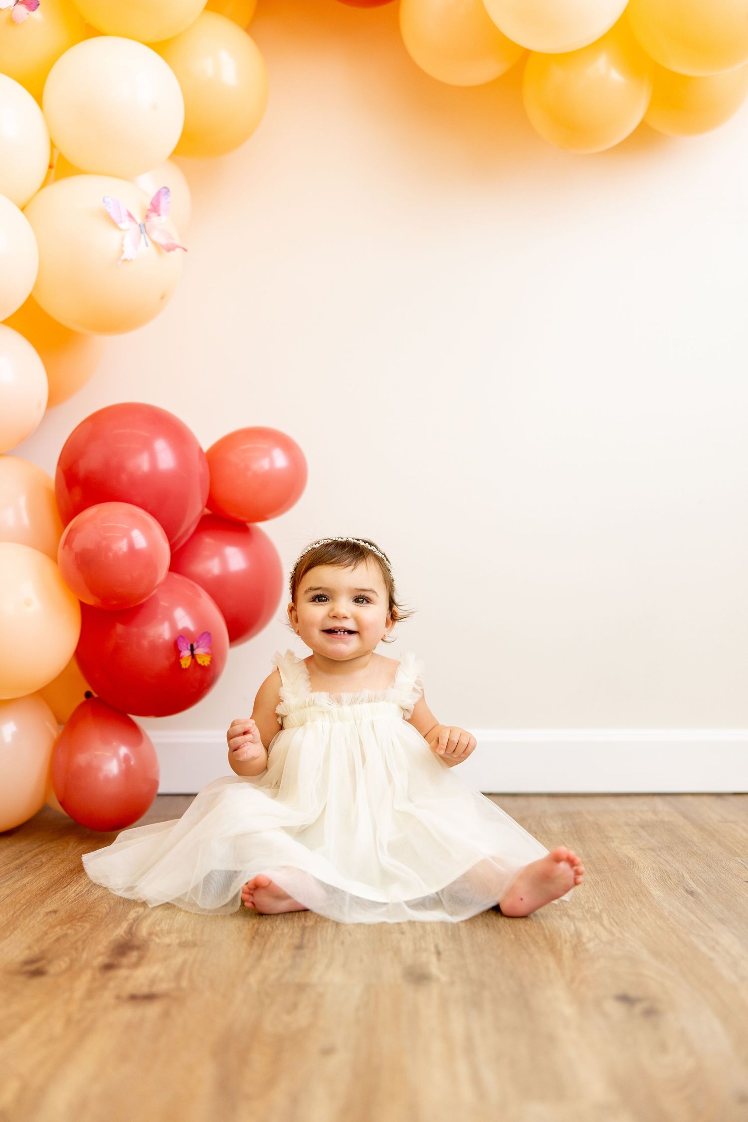 Baby in white dress sitting on hardwood floor with peach and coral balloon arch backdrop.