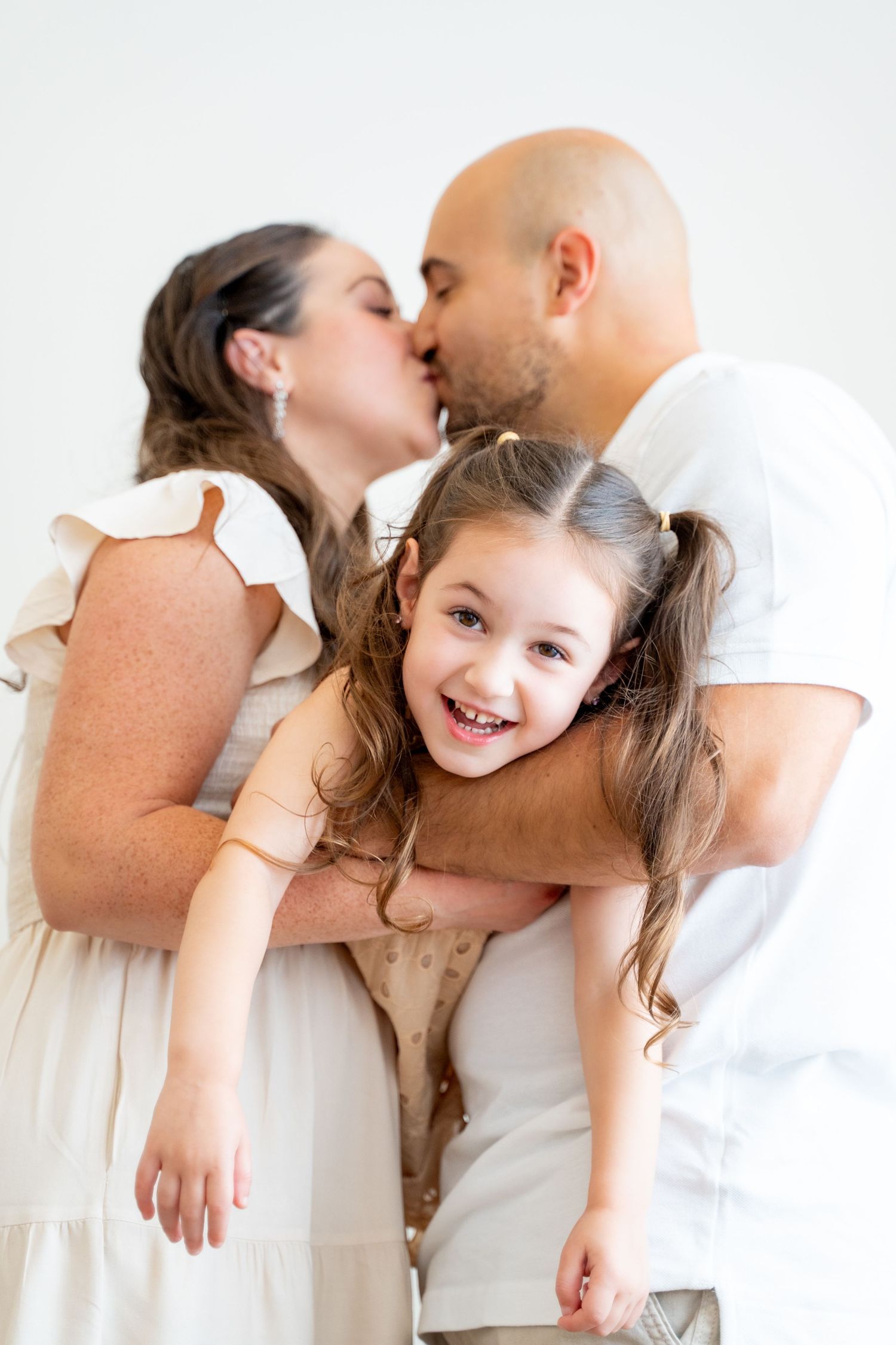 Parents kissing while daughter laughs between them in bright indoor setting.