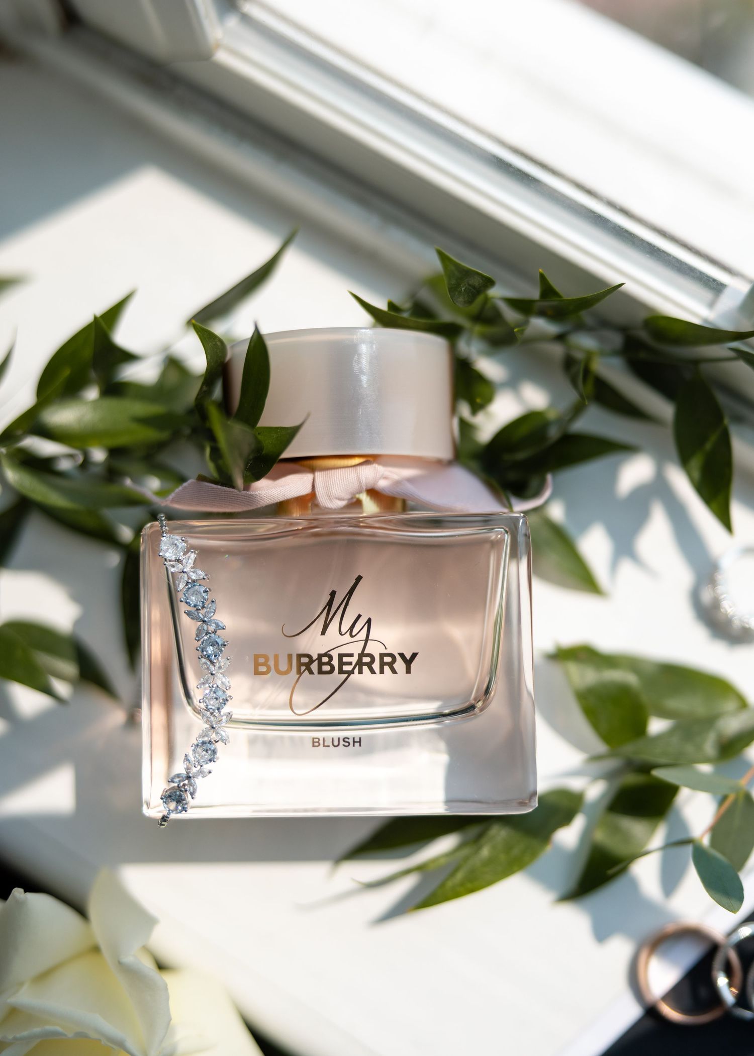 Glass perfume bottle of Burberry fragrance with crystal embellishments surrounded by green leaves in natural light.
