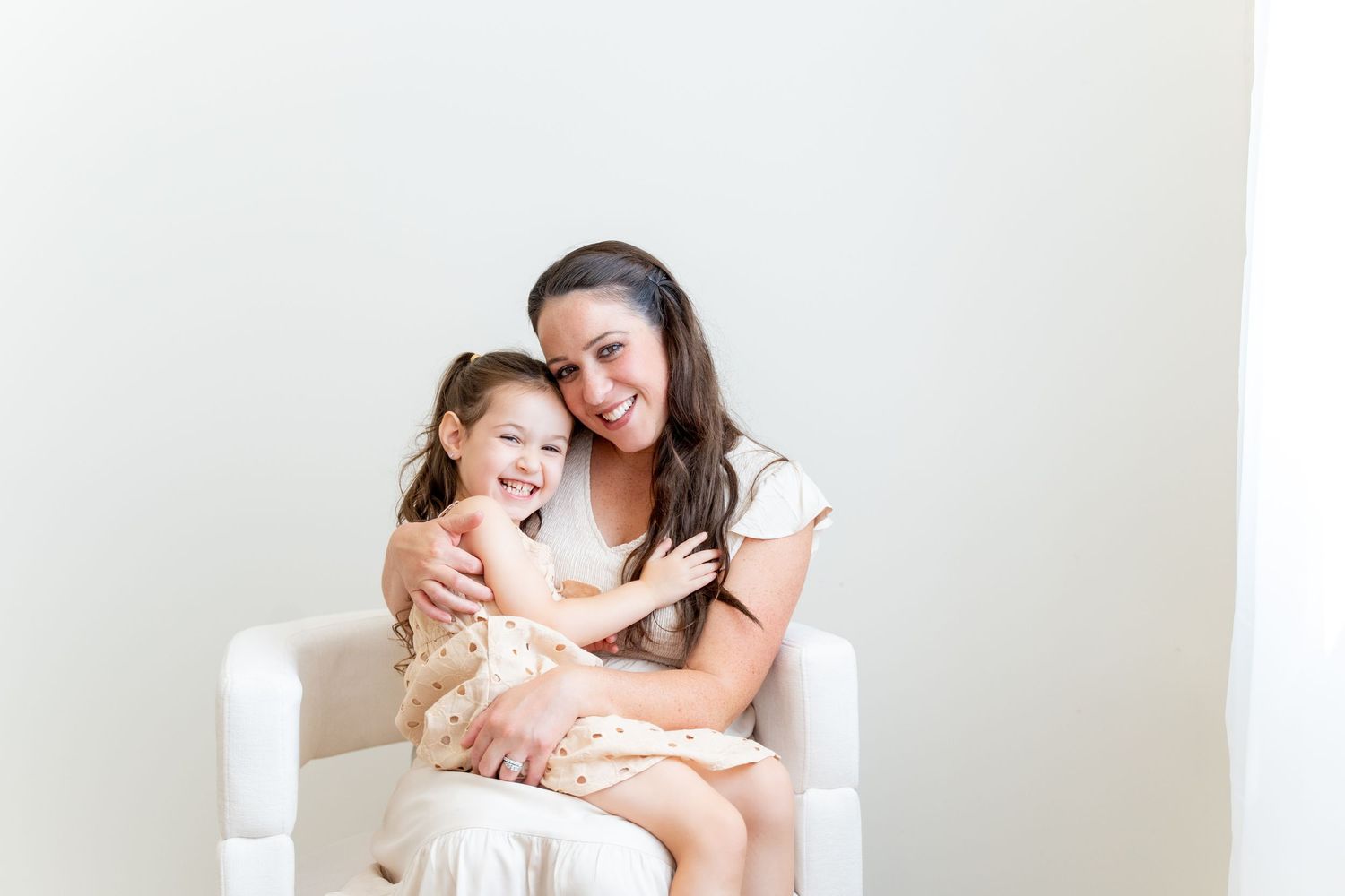 Mother and young daughter sharing joyful moment in white chair against neutral background.