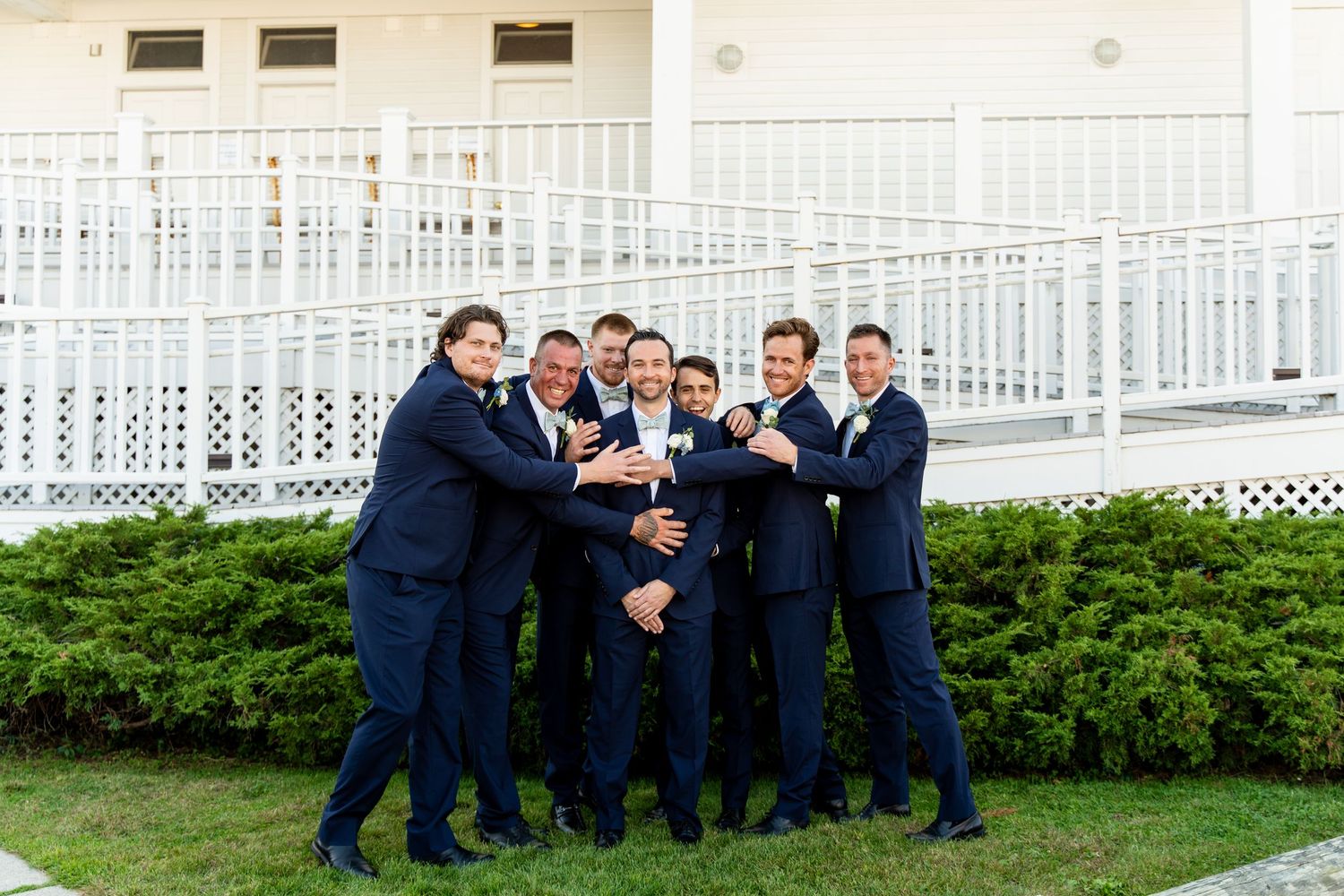 Groomsmen in navy suits gather in playful group pose on lawn in front of white porch.