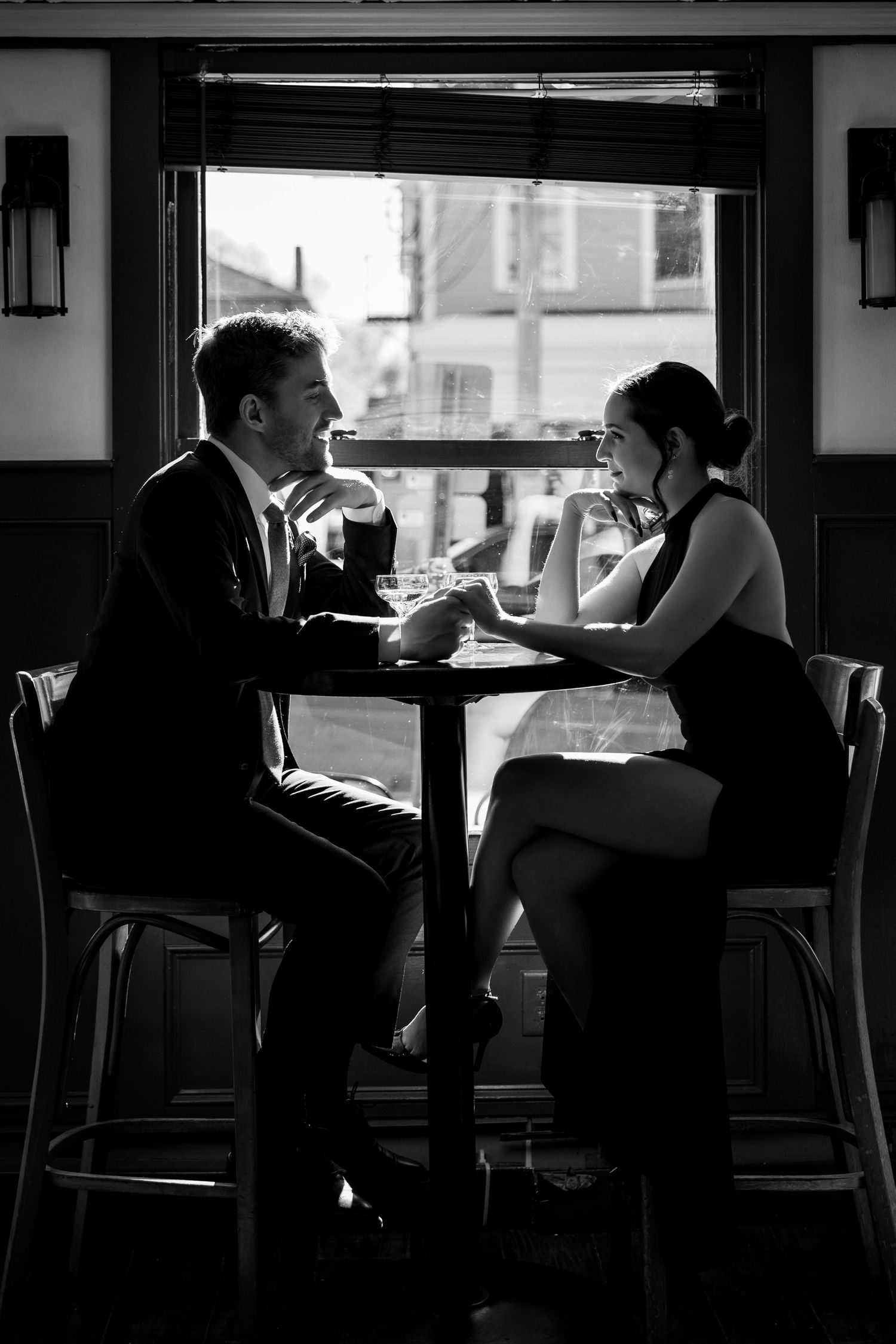 Black and white photo of couple sitting at high-top table near window.