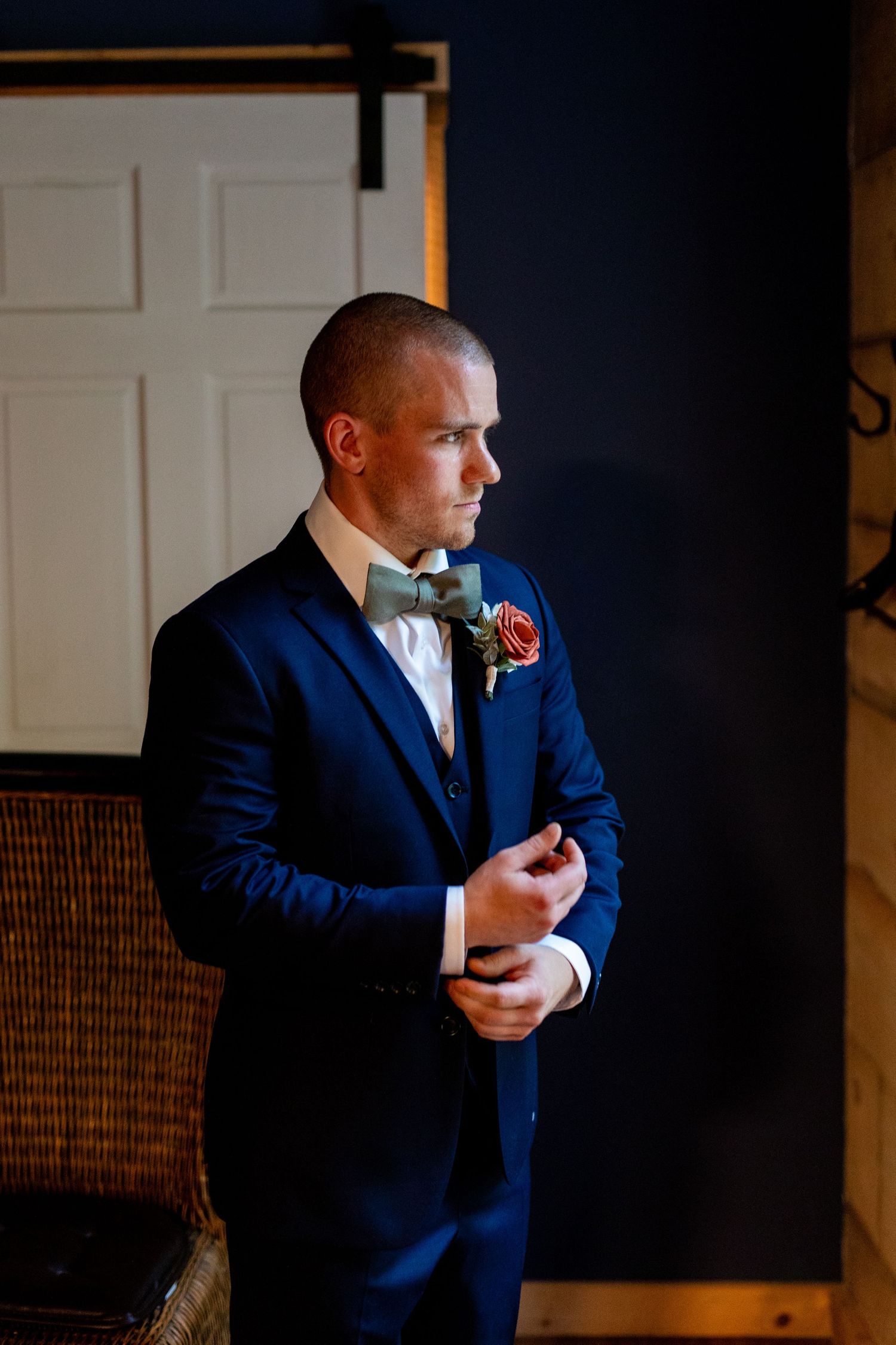 Groom in navy blue tuxedo and bow tie adjusts cuff by window.