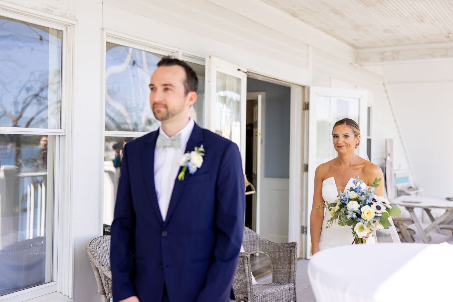 Wedding couple stands on a bright porch with wicker chairs and white decor.