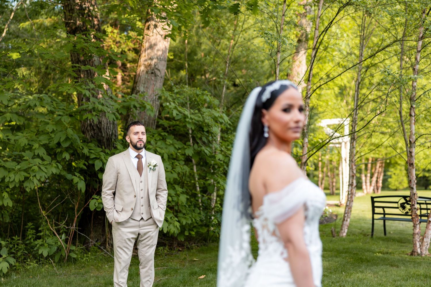 A couple in wedding attire stands in a lush outdoor garden setting with sunlit trees in the background.