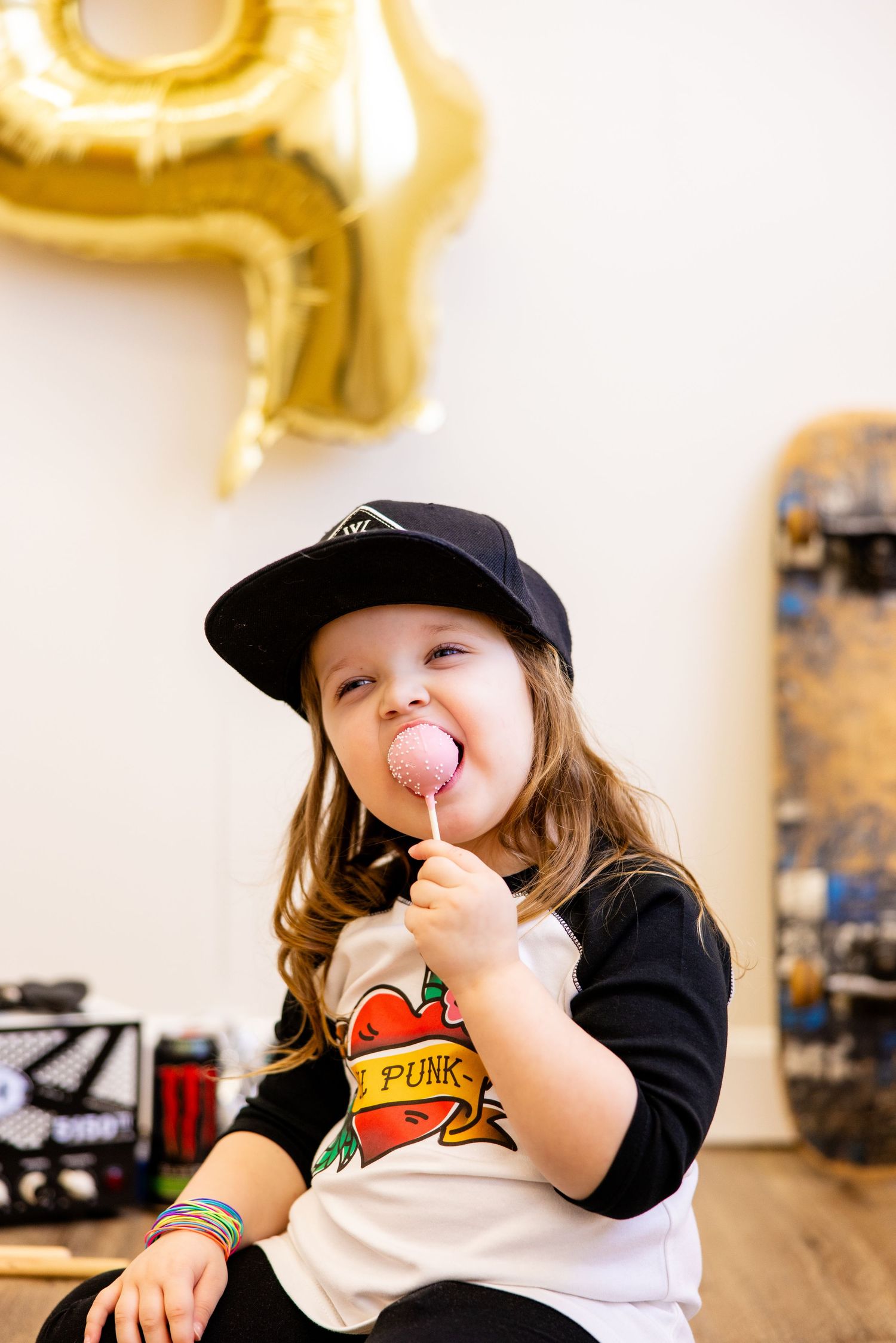 A child wearing a baseball cap enjoys a lollipop while sitting next to skateboarding gear.
