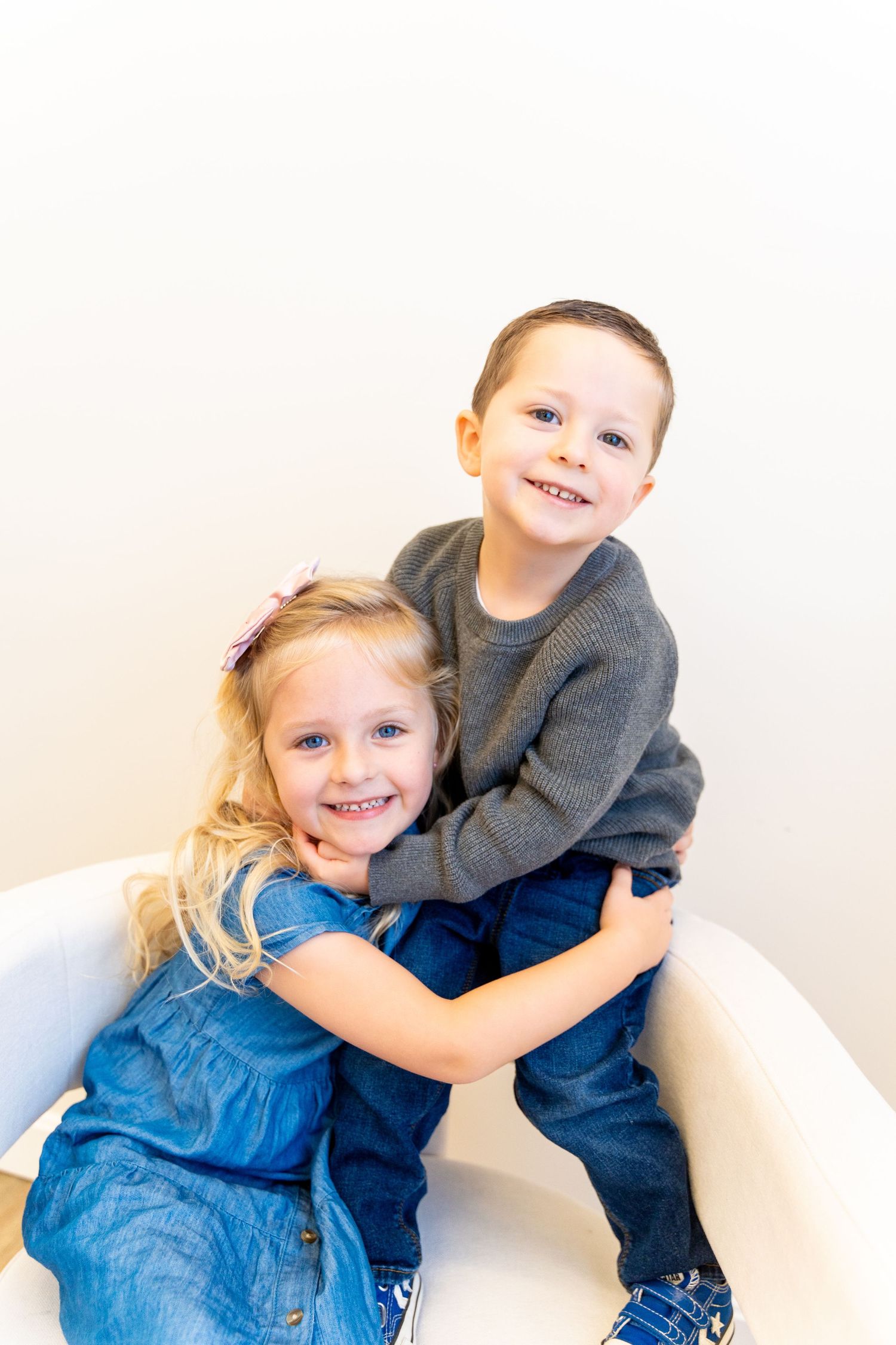 Two children embracing and smiling while sitting on white chair.