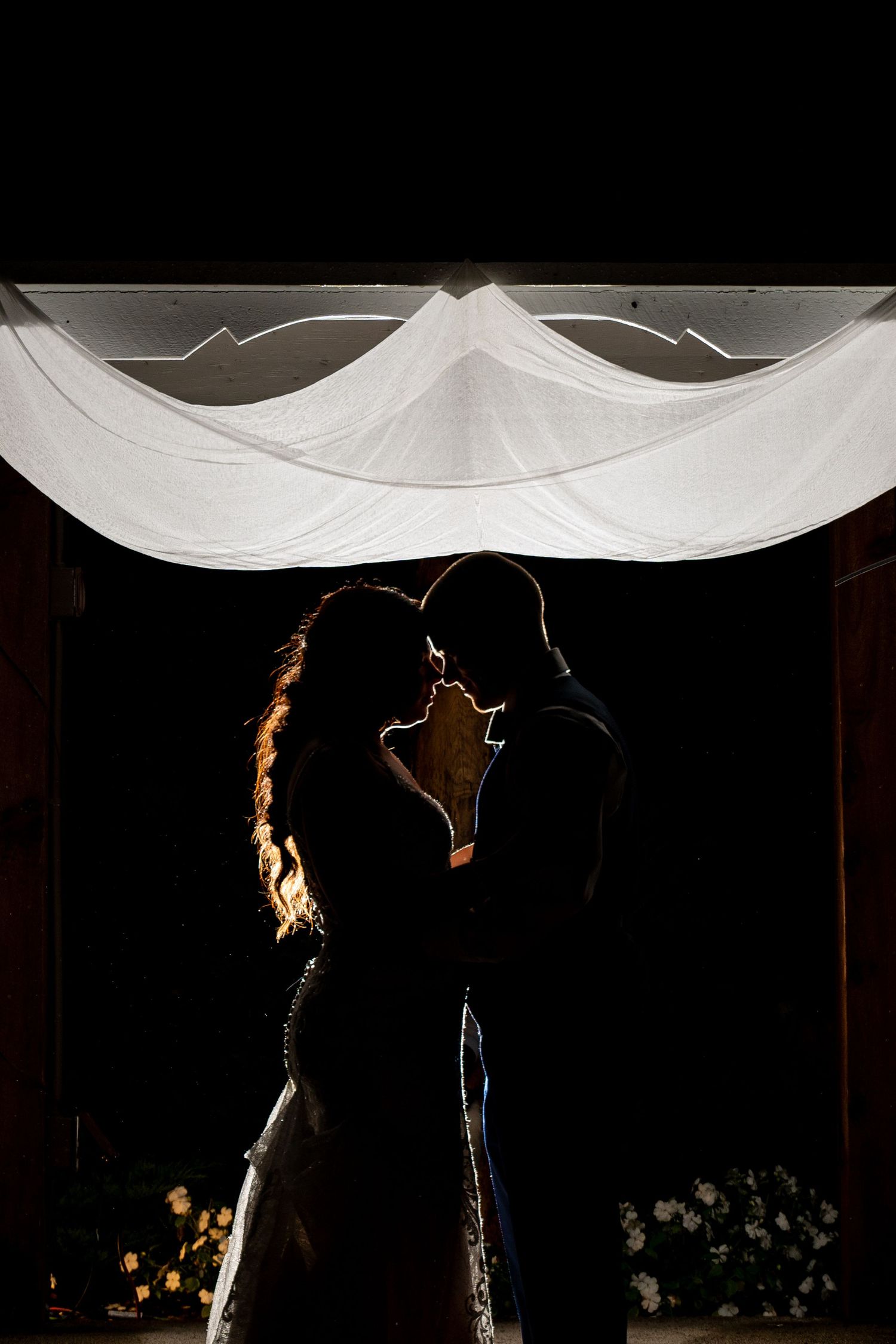 A dramatic backlit silhouette of a couple sharing an intimate moment under white draping.