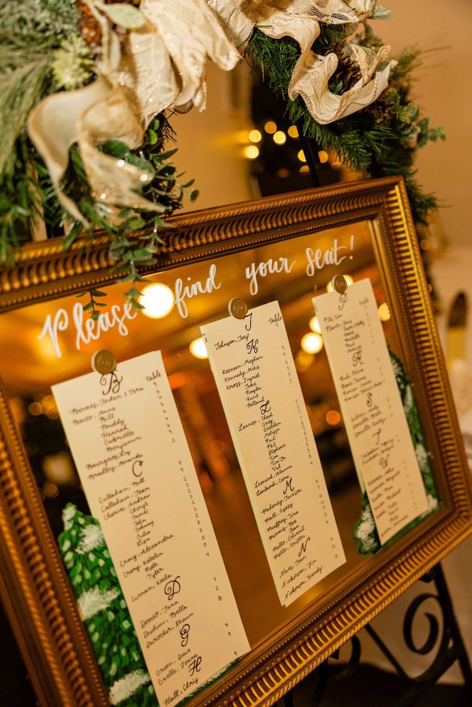 Wedding seating chart displayed in ornate gold frame with string lights and greenery decorations.