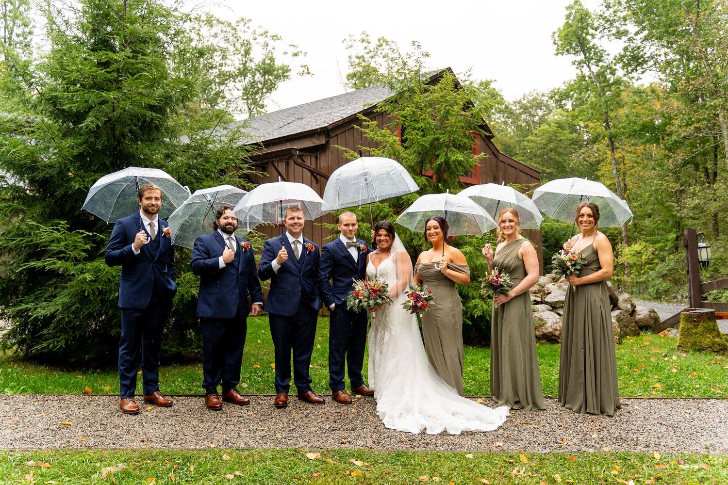 Wedding party poses with umbrellas outdoors wearing navy suits and sage green dresses.