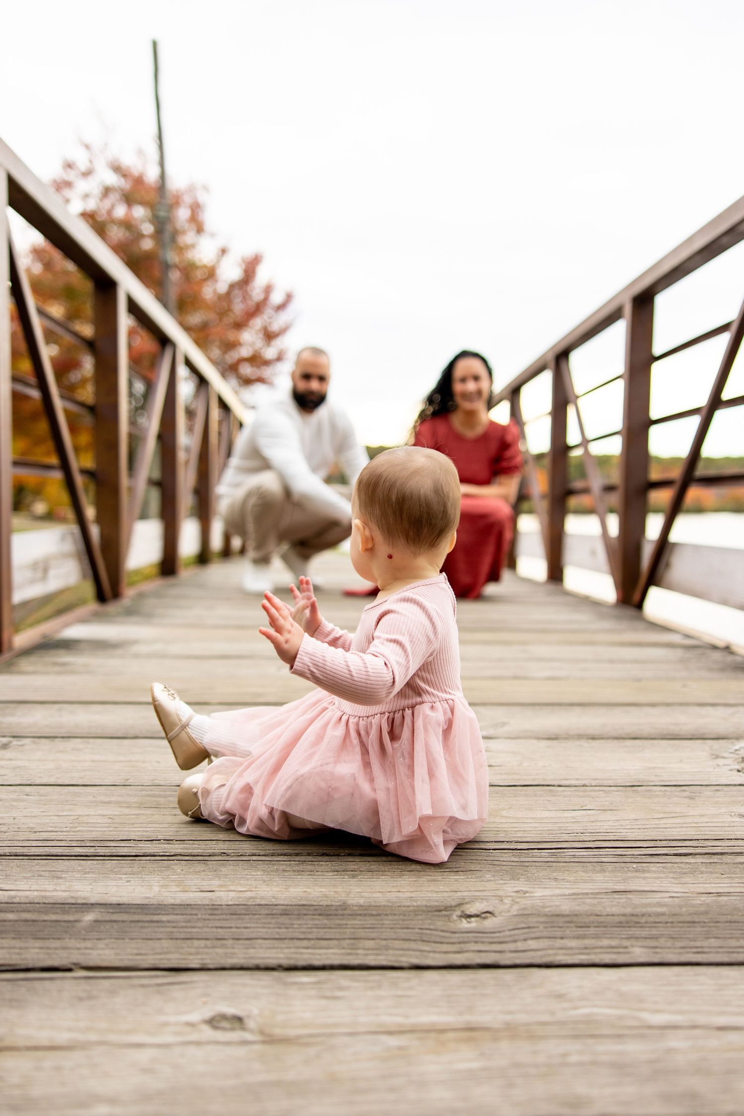 Small child in pink dress sitting on wooden bridge while adults stand in background.