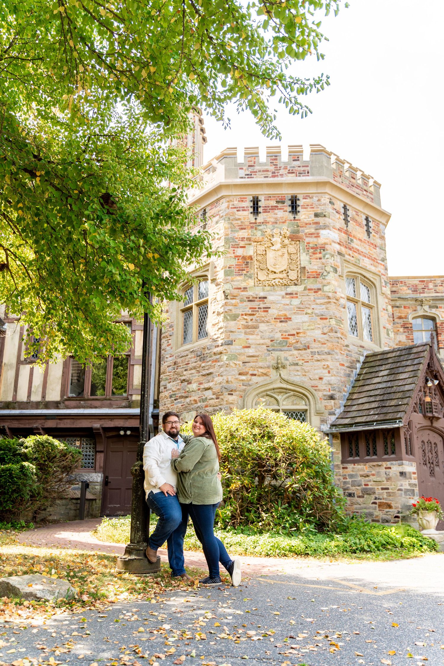 A couple poses together outside a stone castle-like building surrounded by autumn foliage.