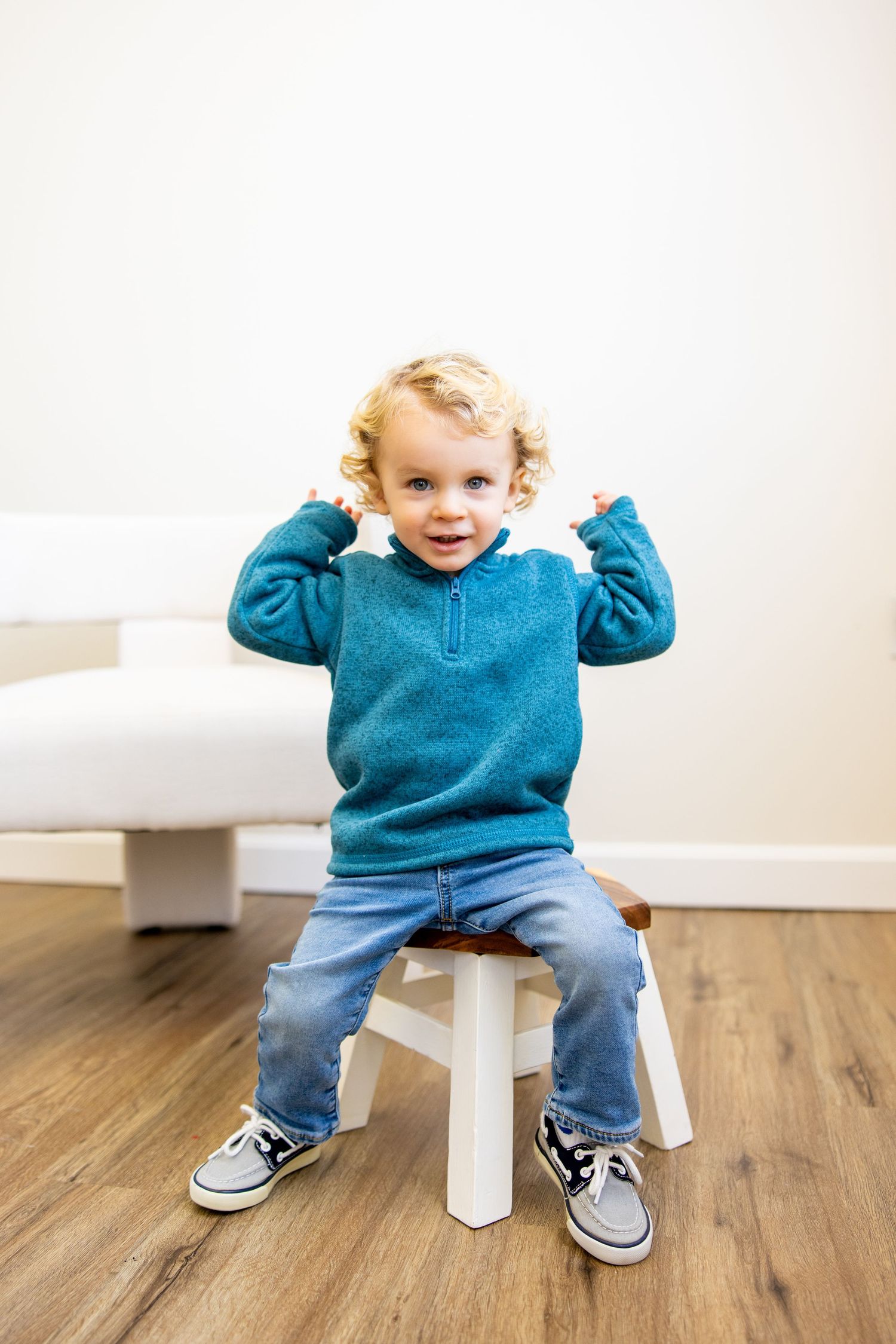 Playful toddler in teal sweater sitting on white stool with arms raised.