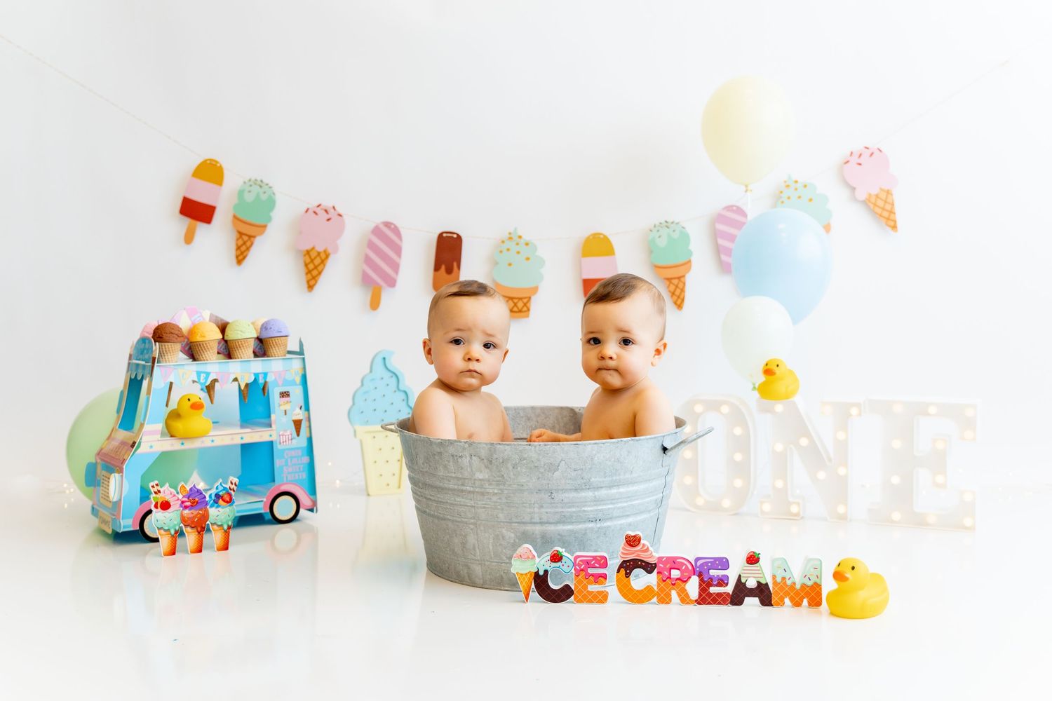 First birthday celebration setup with ice cream themed decorations, twin babies sitting in a metal tub.