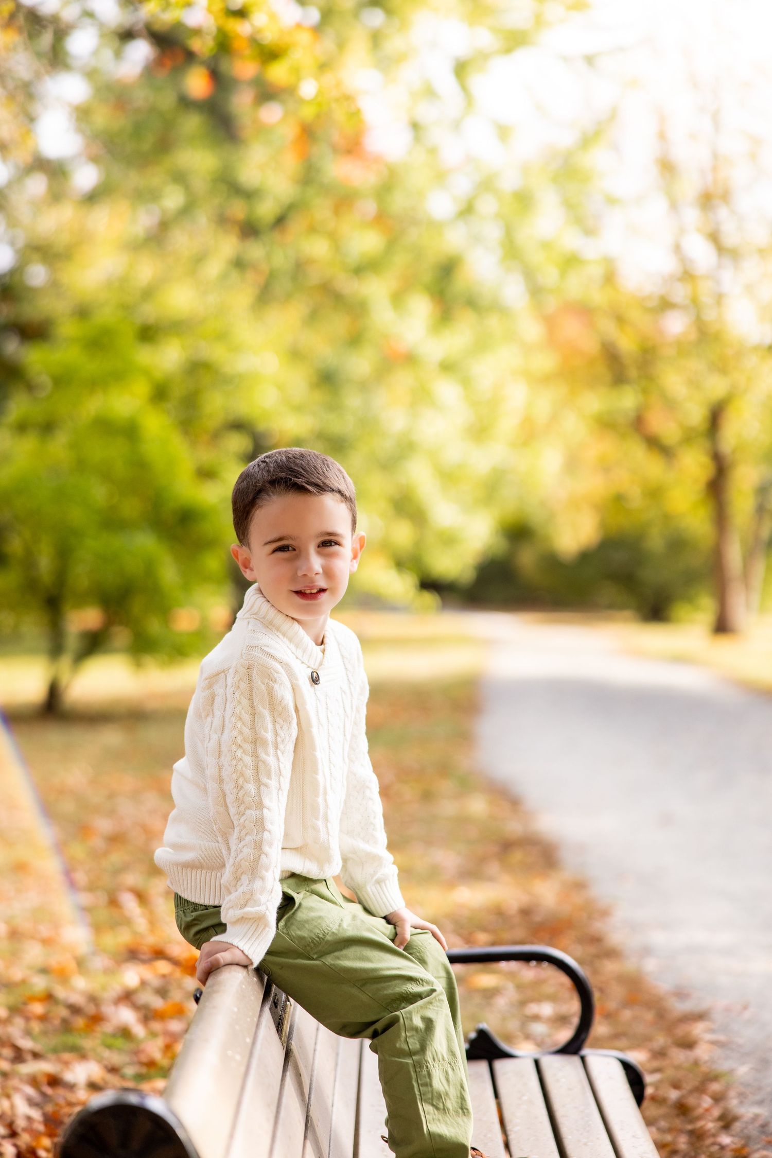 Child in white cable knit sweater sitting on park bench surrounded by fall foliage.