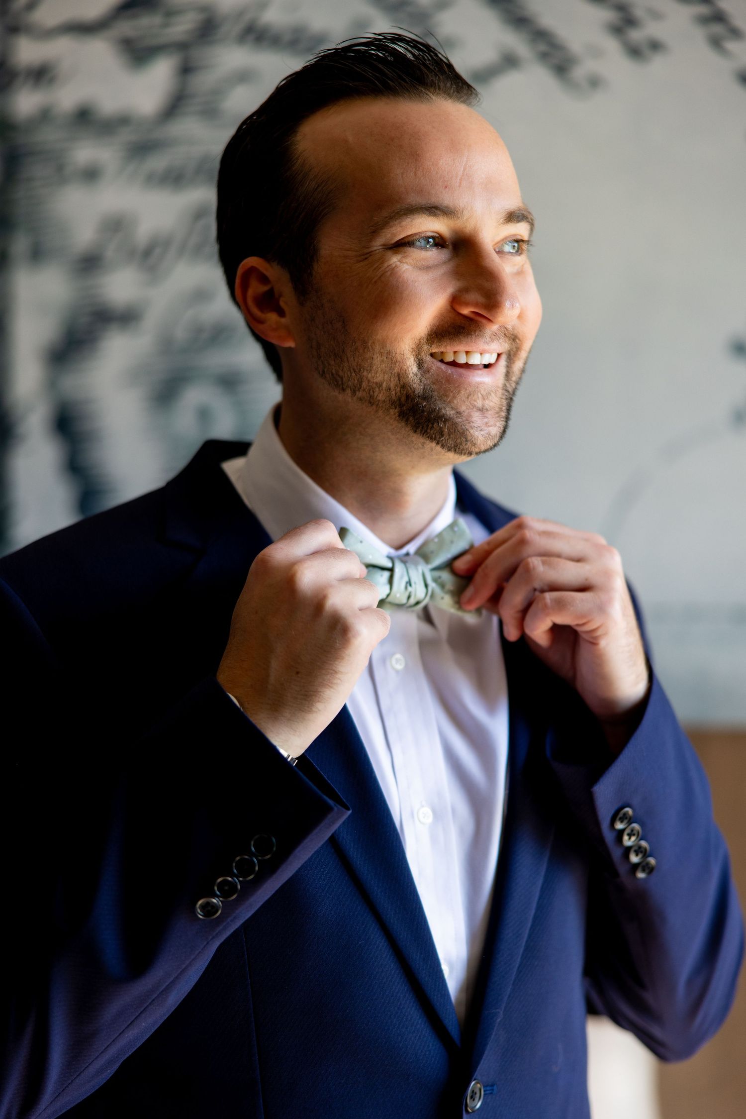 Groom adjusts mint green bowtie while wearing navy suit and white shirt in preparation for wedding day.