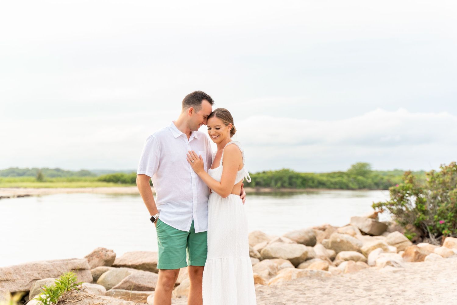 A romantic couple embraces on a rocky beach with water and cloudy skies in the background.